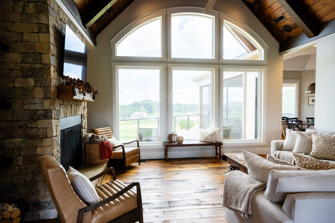 Living room with large arched windows, stone fireplace, hardwood floors, and beige and white couches.