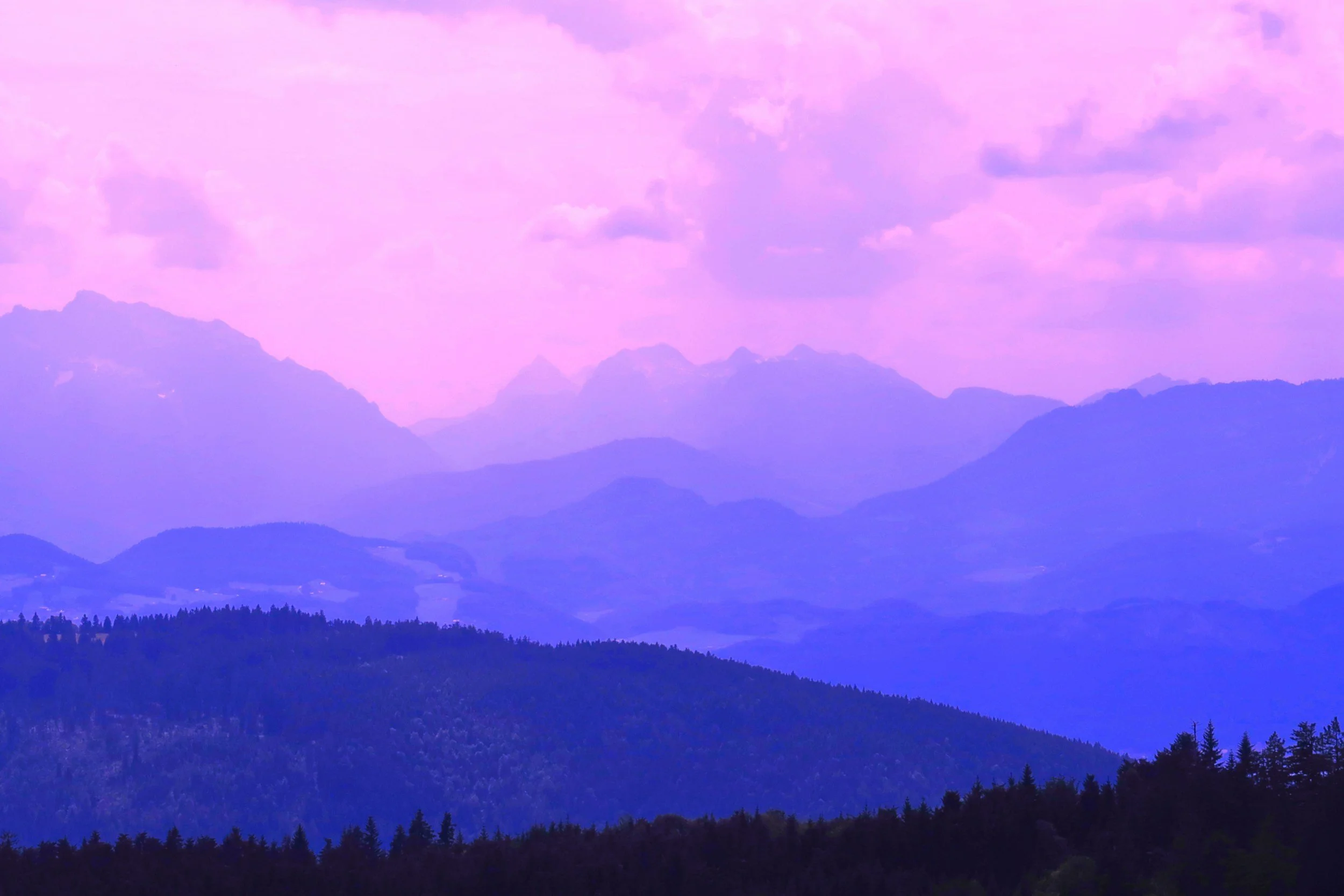 A scenic view of mountain ranges and hills at dusk with pink and purple hues in the sky, and dark forested areas in the foreground.