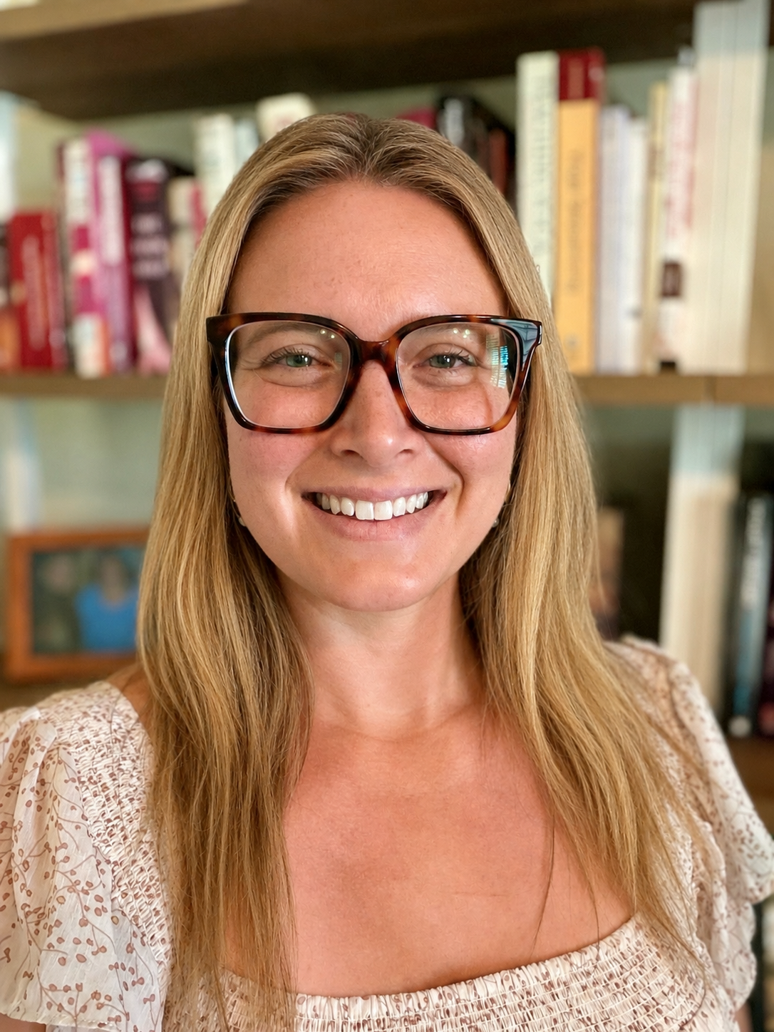 A woman with long blonde hair wearing large tortoiseshell glasses smiling in front of a bookshelf filled with books.