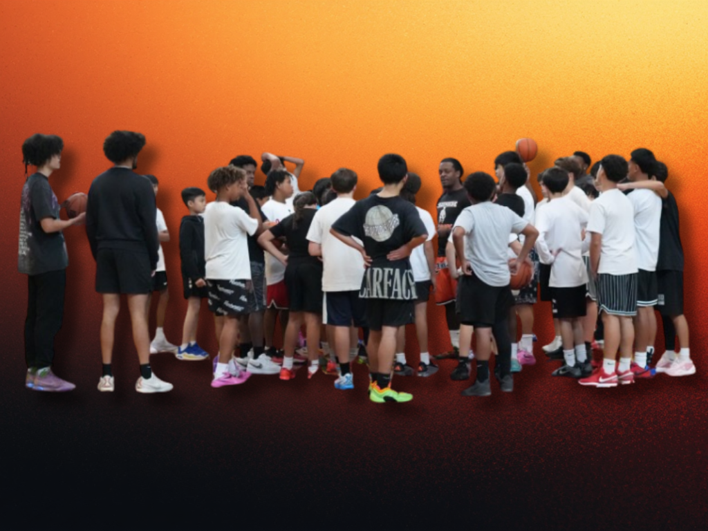 A group of children and teenagers gathered around a person giving a basketball lesson in a gym or sports facility with a painted orange wall.
