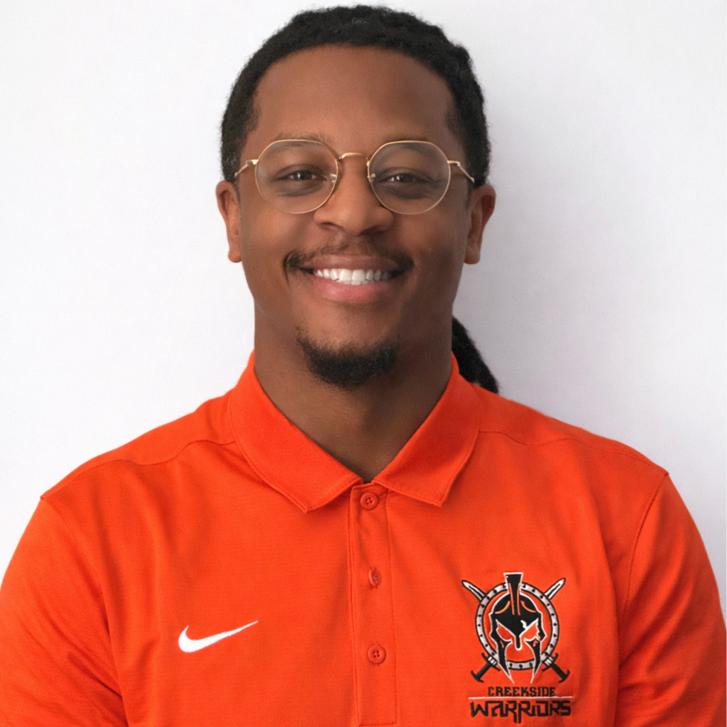 A young man smiling, wearing glasses and an orange polo shirt with a Creeksde Warriors logo.