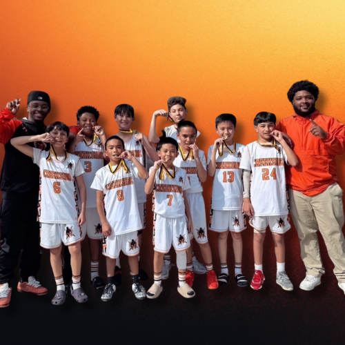 Group of young boys in basketball uniforms with medals, posing with two coaches or teachers, against a vibrant orange gradient background.