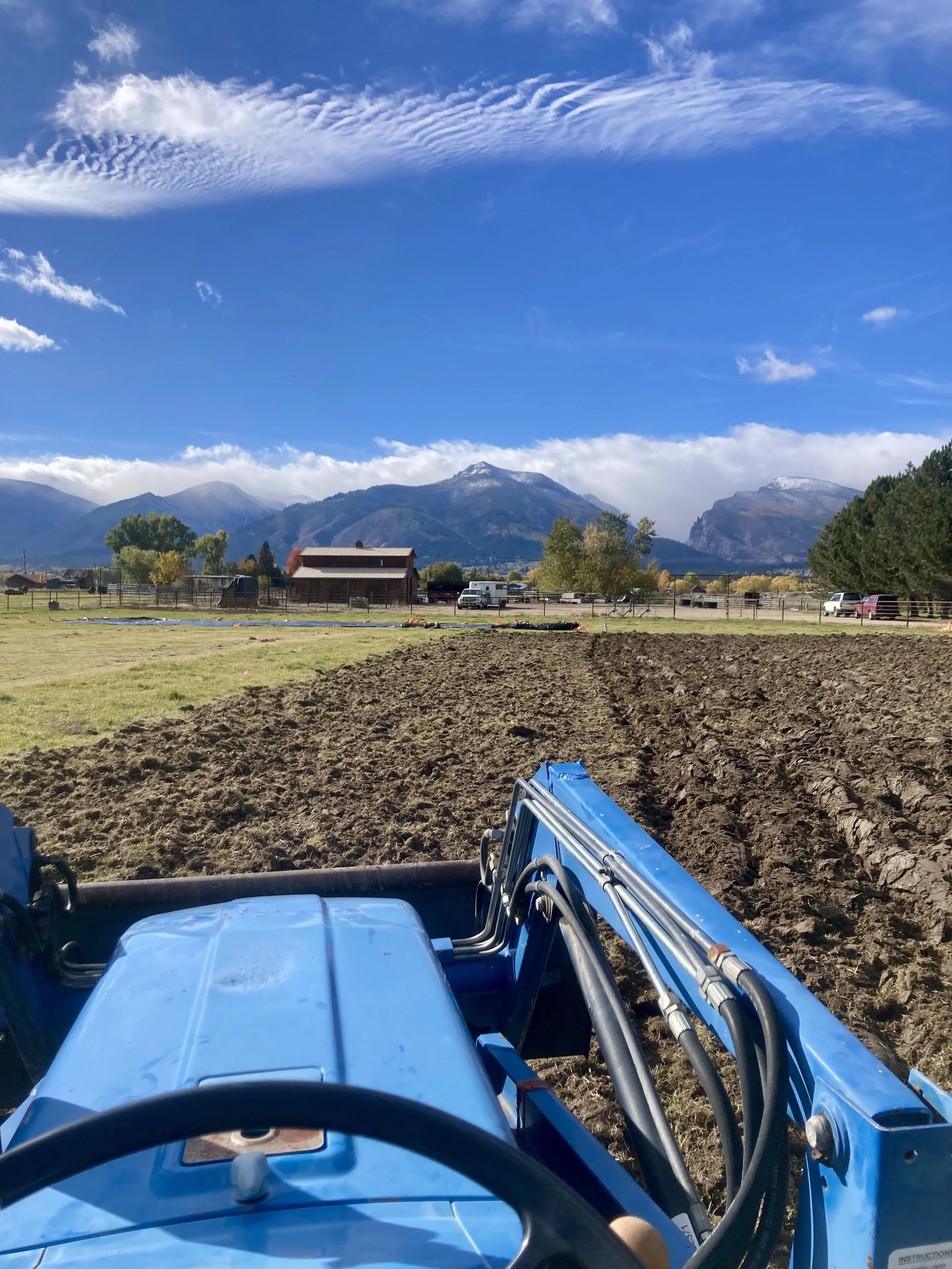 View from a tractor seat showing freshly plowed soil on a field, with mountains and blue sky with clouds in the background.