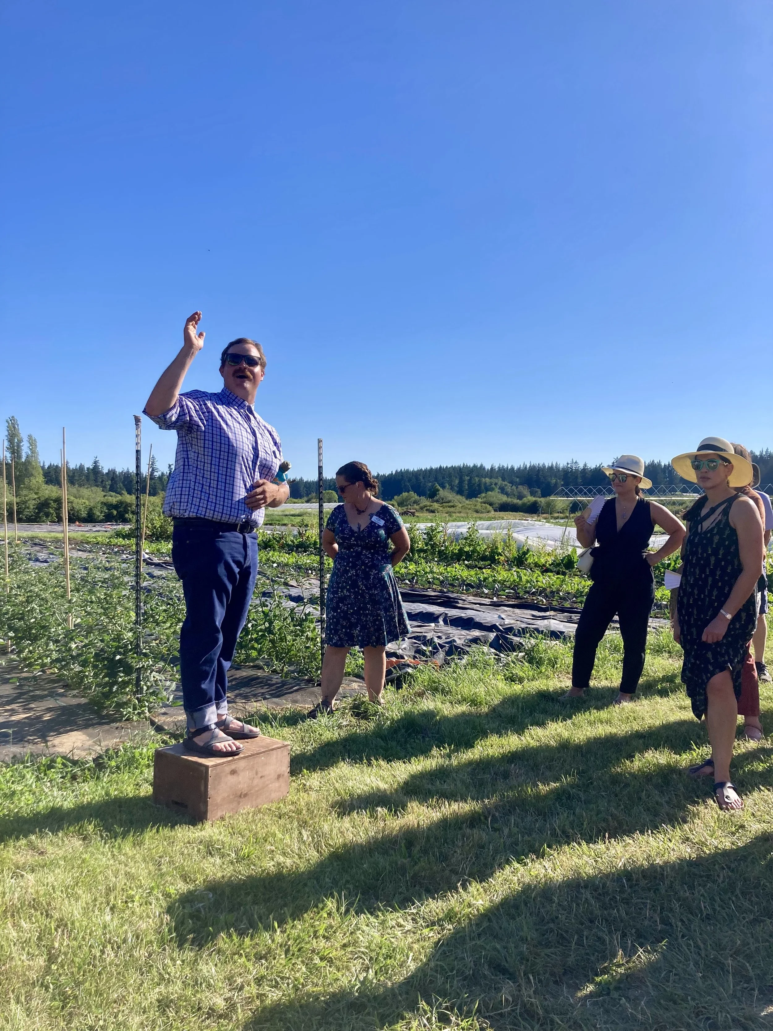 A man standing on a wooden block at a farm, speaking to a group of women wearing summer dresses and hats on a sunny day.