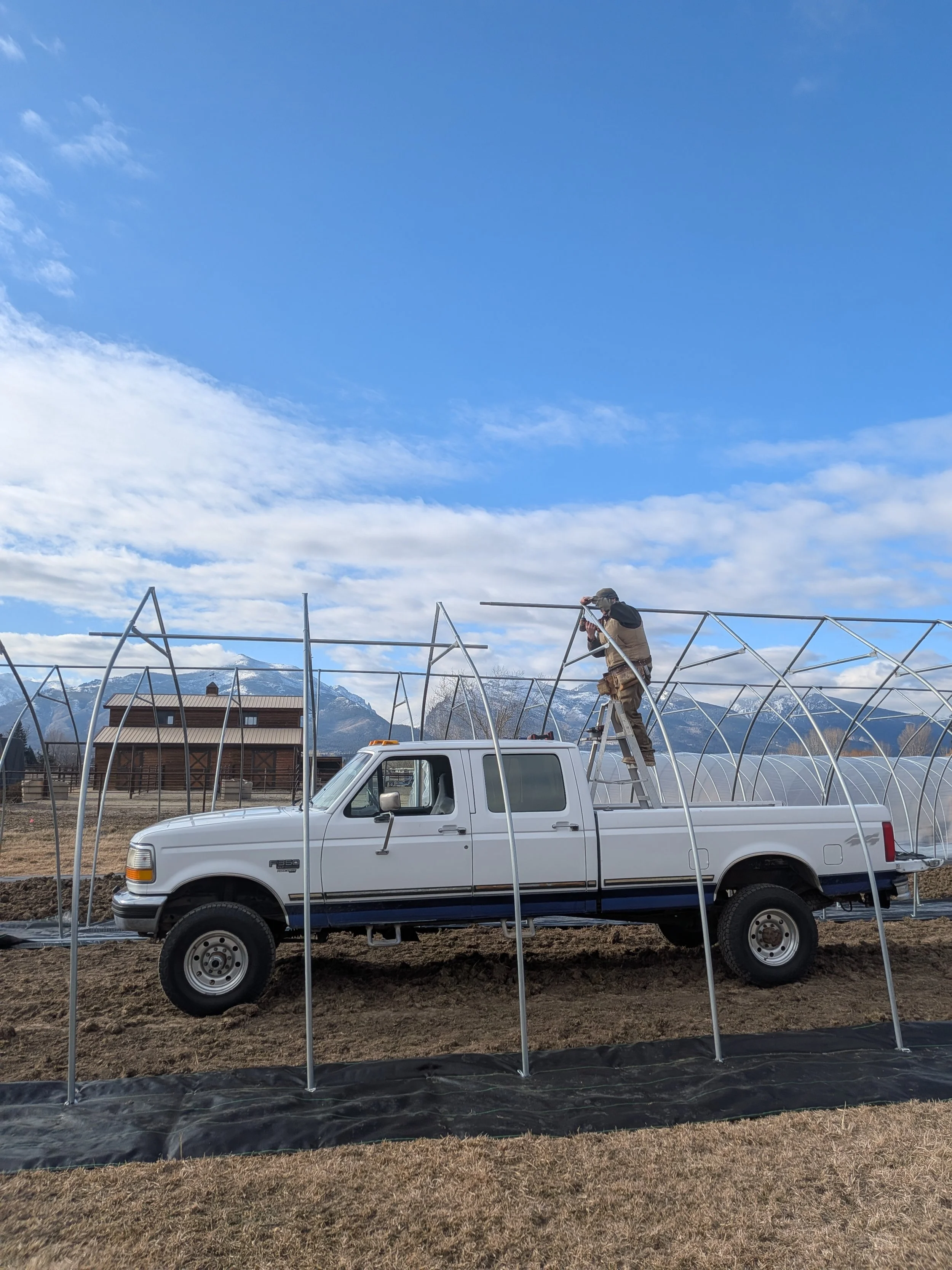 A person working on metal greenhouse framework mounted on a white pickup truck in a rural area with mountains and blue sky in the background.