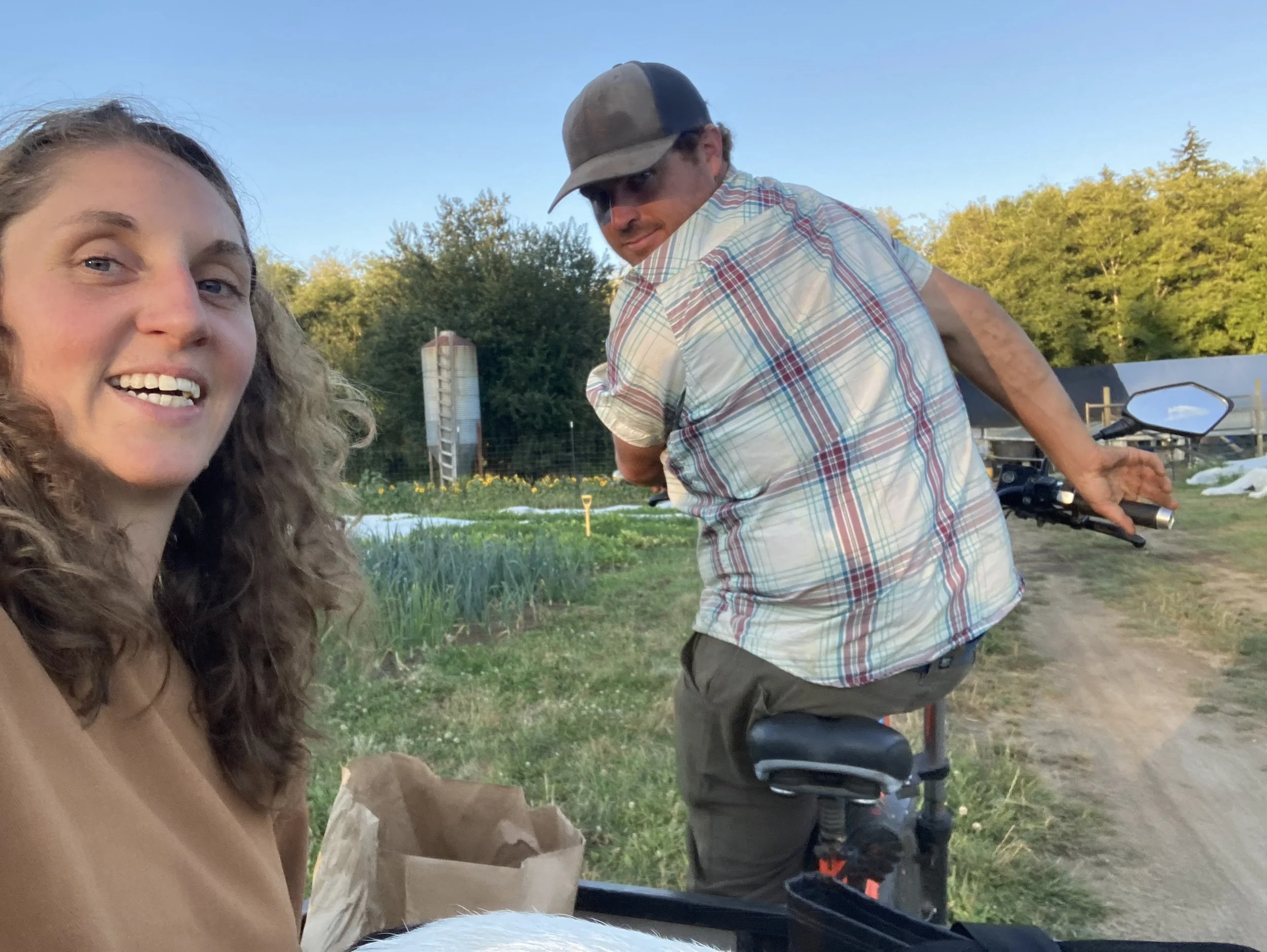 A woman taking a selfie with a man on a bicycle in a rural outdoor setting during daytime. The woman has long, curly hair and is smiling at the camera. The man is wearing a plaid shirt and a cap, looking toward the camera, and holding a bicycle.
