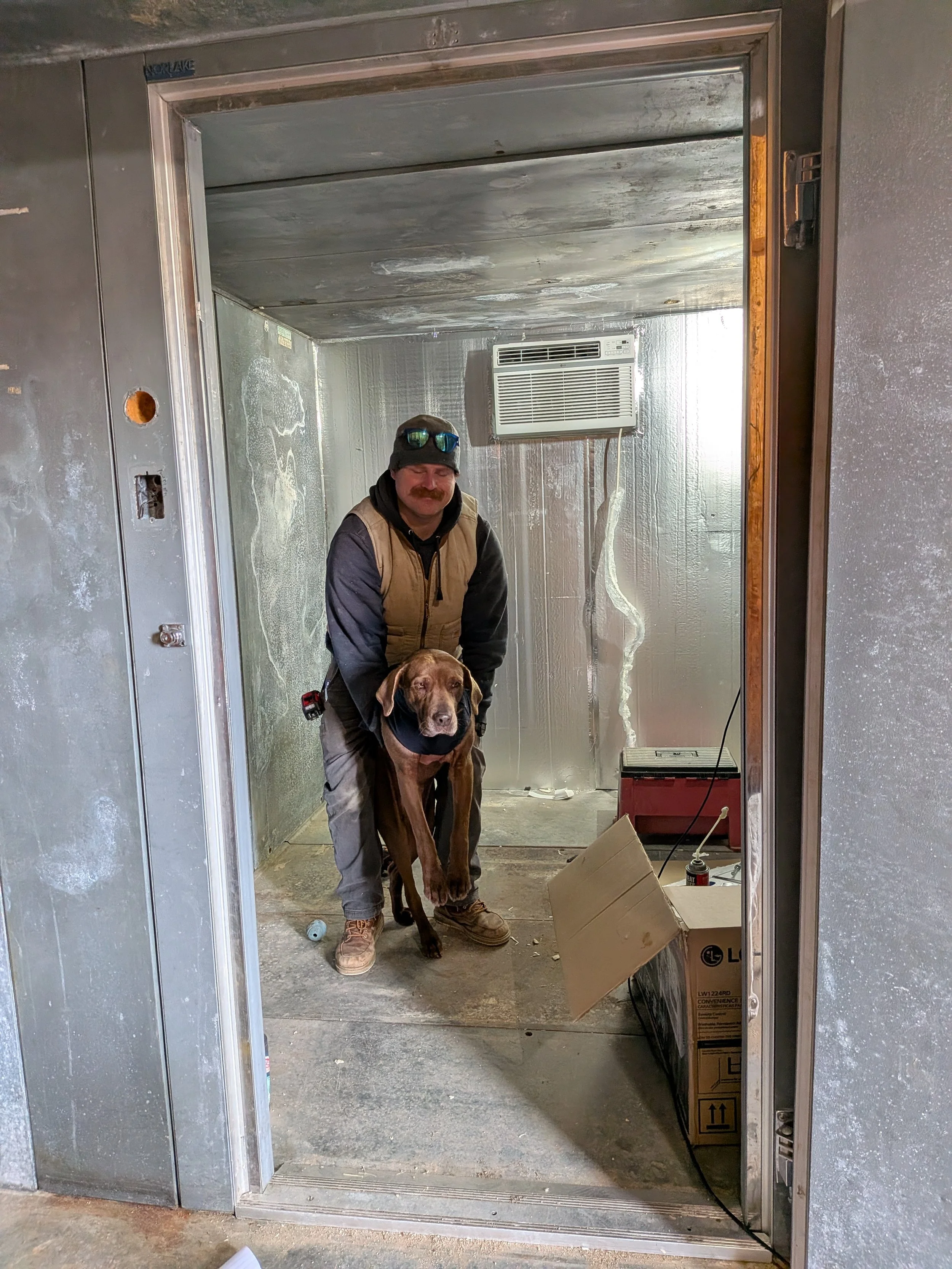 A man and a brown dog standing inside a walk-in fridge, holding a dog by the front legs.