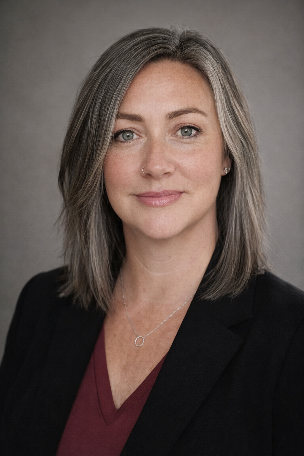 Professional headshot of a woman with shoulder-length gray hair, wearing a black blazer and maroon top, smiling softly, against a gray background.