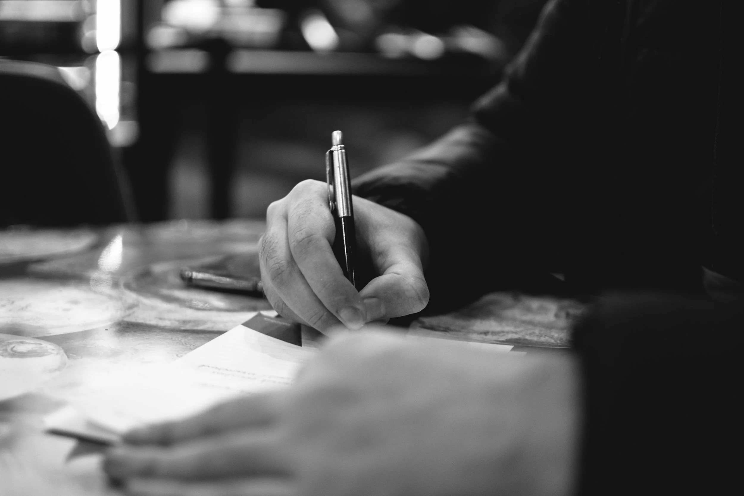A person writing with a pen at a table, black and white photo focusing on their hand and the pen.