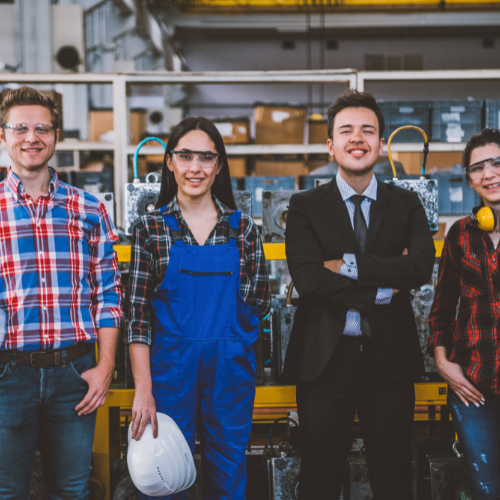 Four young adults, two men and two women, standing together in an industrial or workshop setting with equipment and shelves in the background. They are smiling and appear to be colleagues or students.