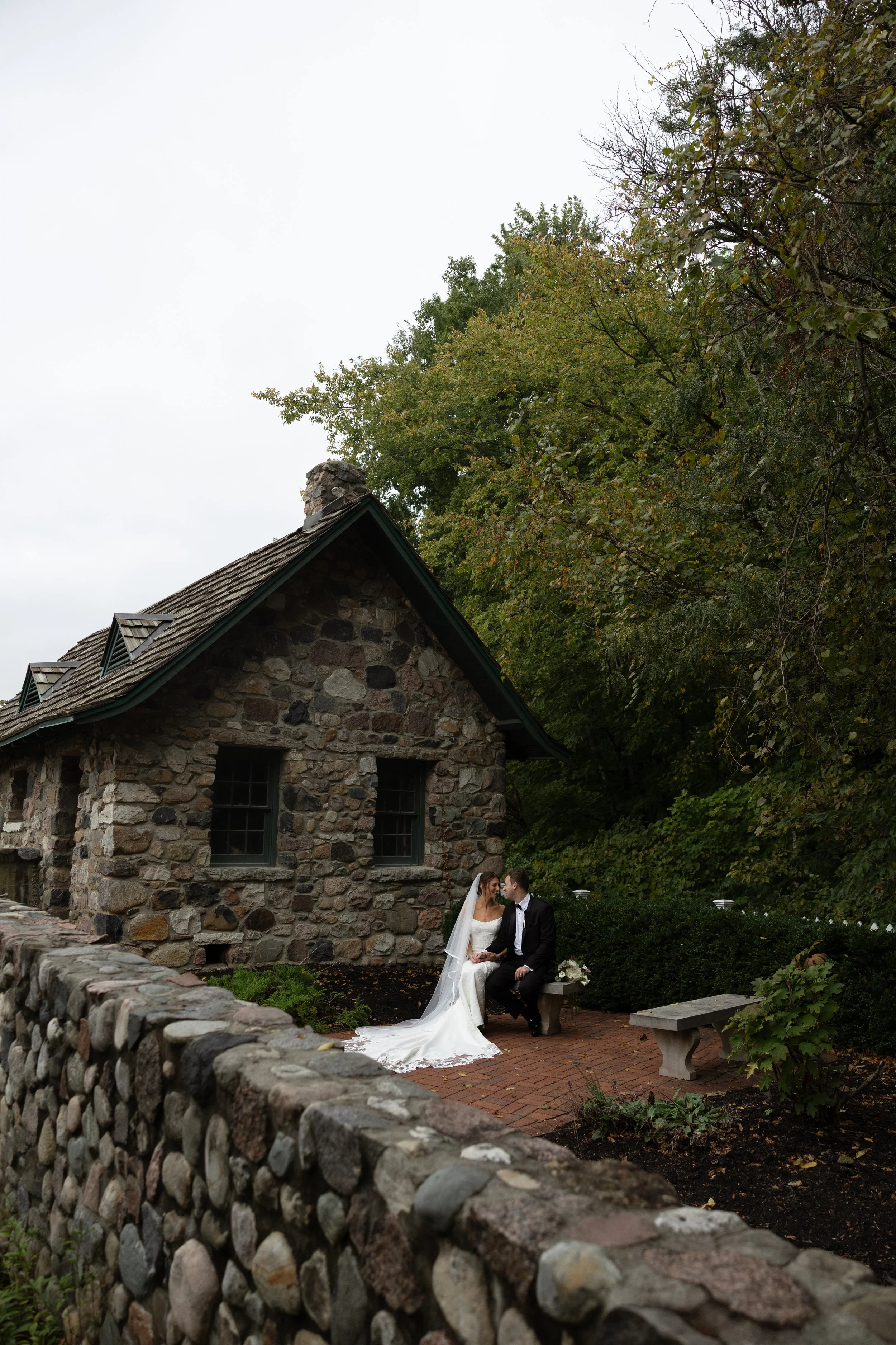 Bride and groom seated outside a rustic stone cottage surrounded by trees and garden landscaping