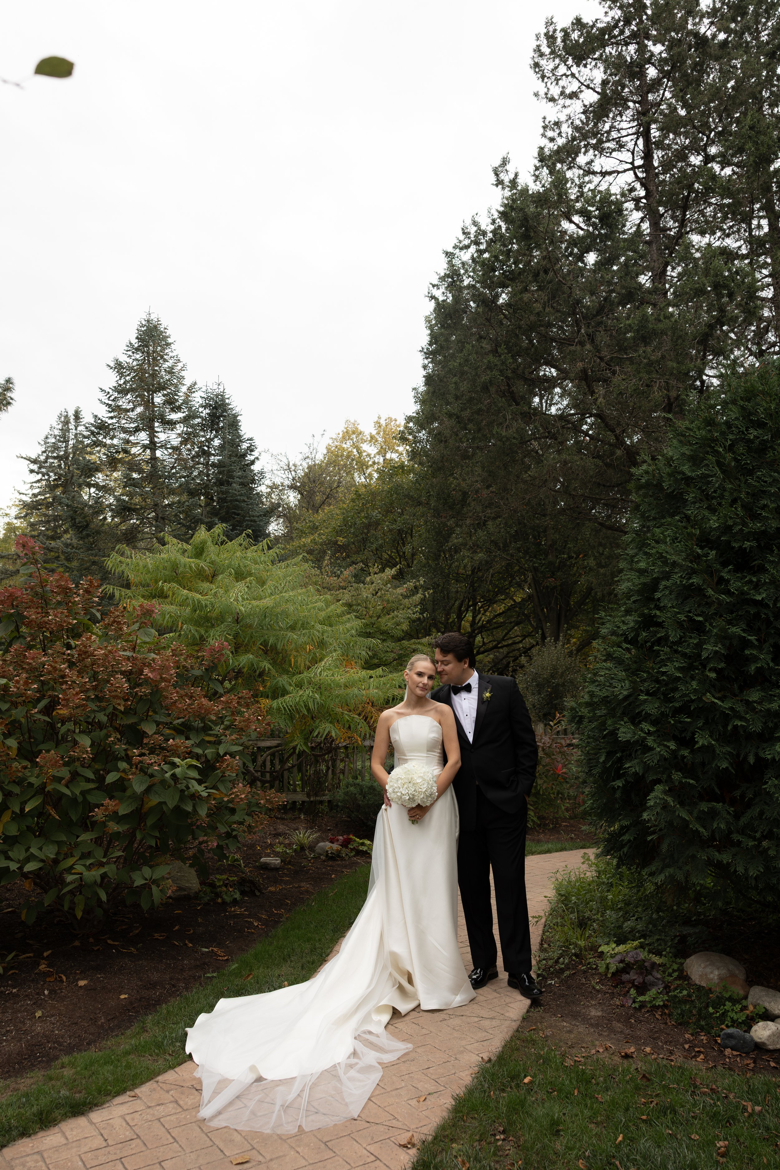 Bride and groom posing with a white bouquet on a garden path surrounded by trees