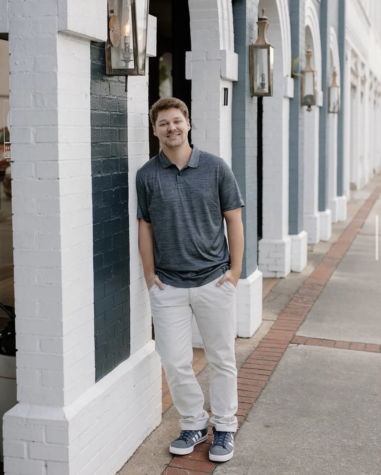 A young man with brown hair, wearing a dark gray polo shirt, white pants, and blue sneakers, smiling while standing against a white and dark blue brick wall on a city sidewalk.