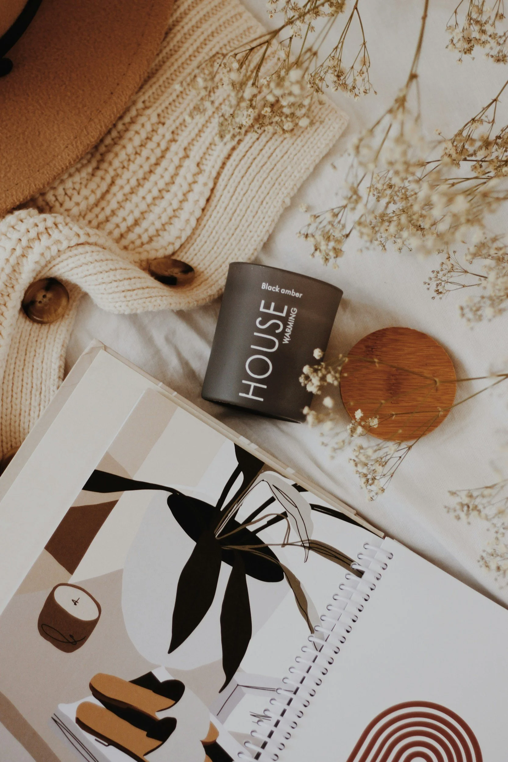 A beige knitted cardigan with wooden buttons, a black mug labeled 'HOU$E' with white text, a small wooden circular dish, white dried flowers, a spiral-bound notebook with abstract plant artwork, and a beige background.