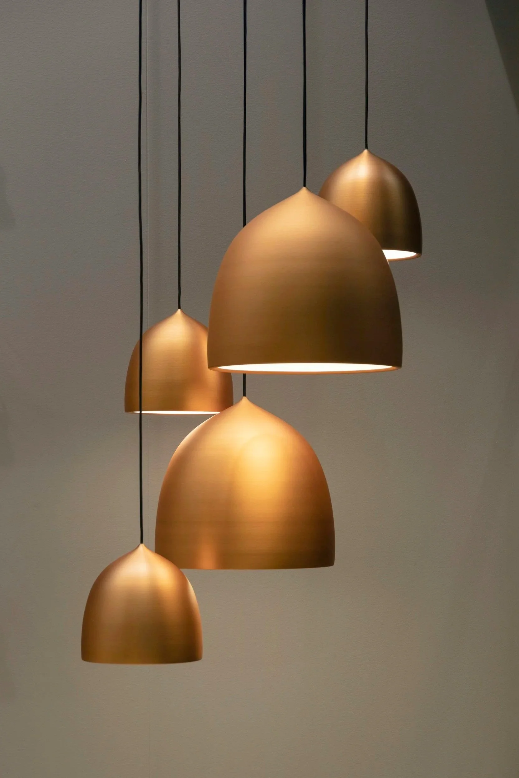 A modern chandelier with five gold-colored dome-shaped pendant lights hanging from black cords against a plain wall.