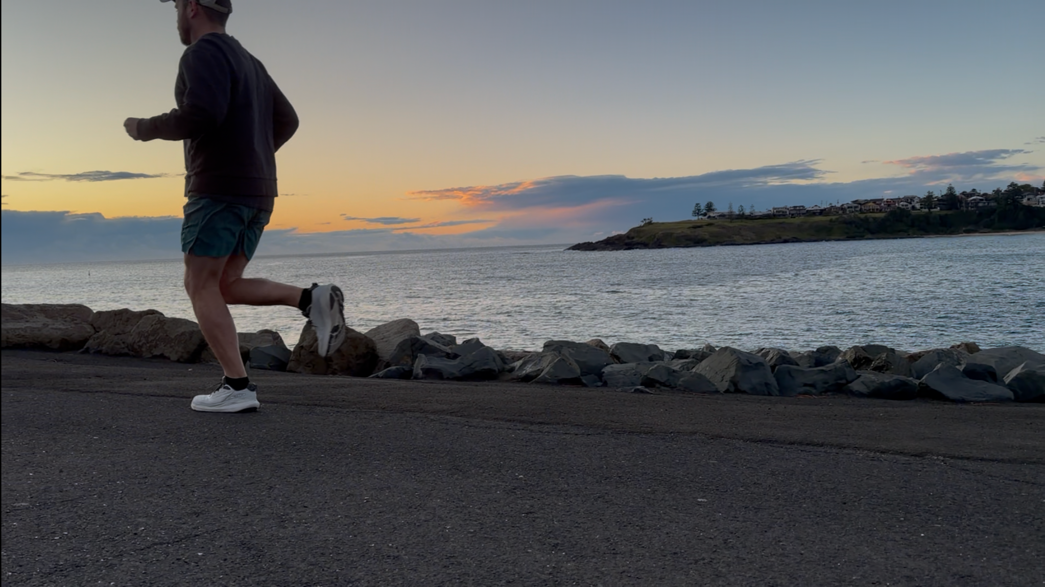 A person jogging along a rocky shoreline at sunset, with water and a distant hillside with houses in the background.