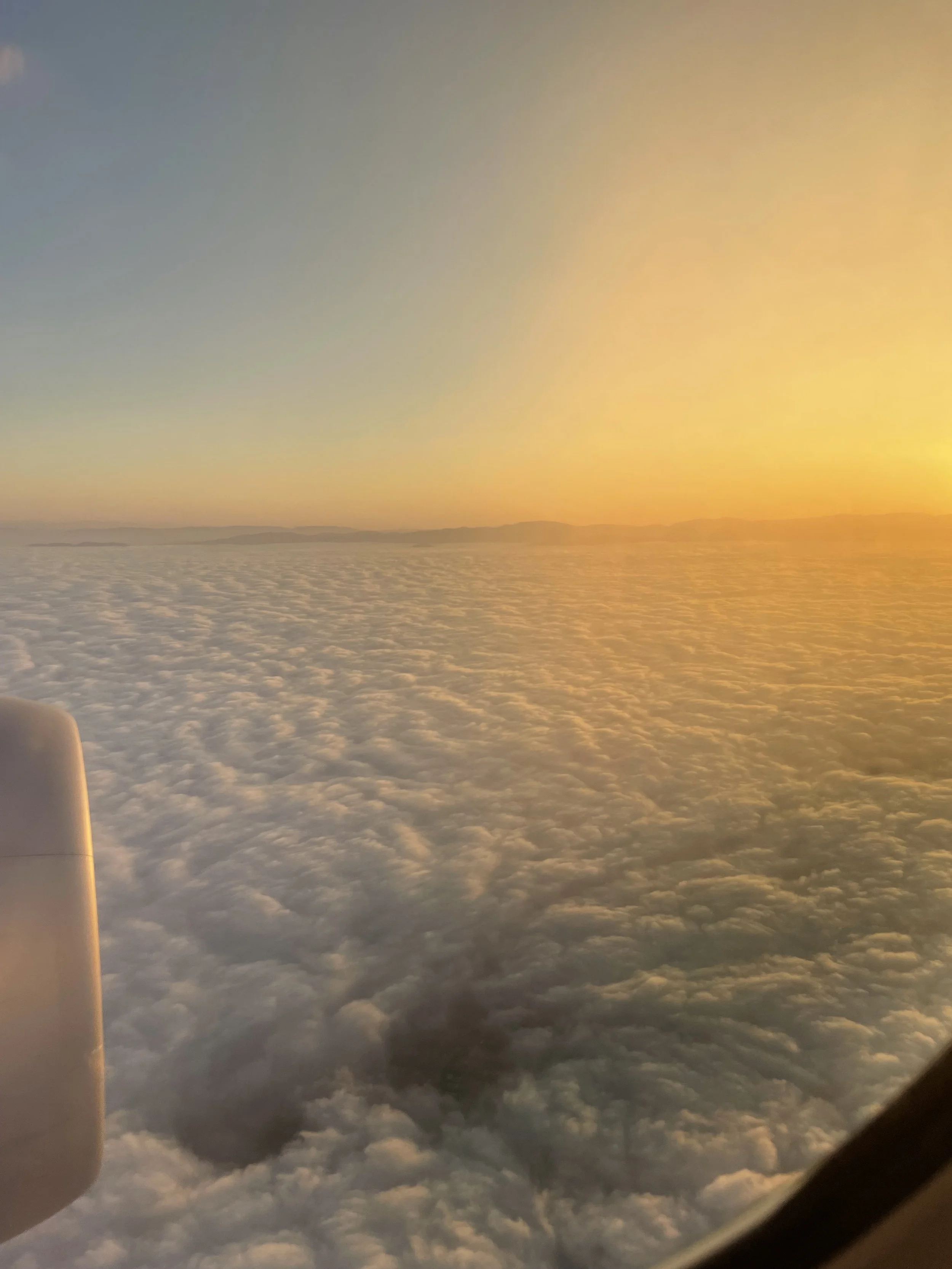Image looking outside of an airplane window. Jet engine is visible. There is an orange sunrise and mountains breaking above the clouds.