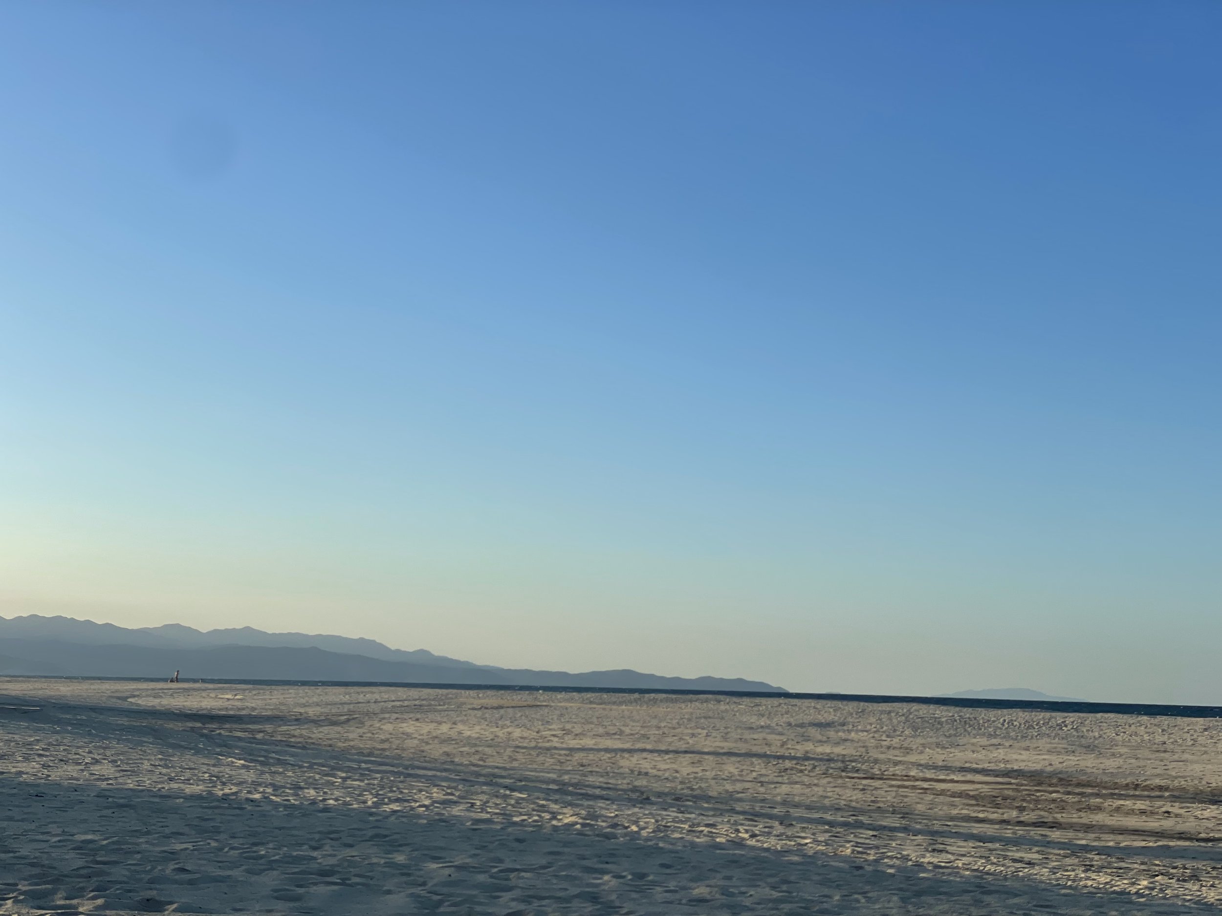 Photo is of a sandy beach on the Four Seasons Los Cabos property at sunset. There are footprints in the sand and the sun behind the mountains on the edge of the Four Seasons Los Cabos property.