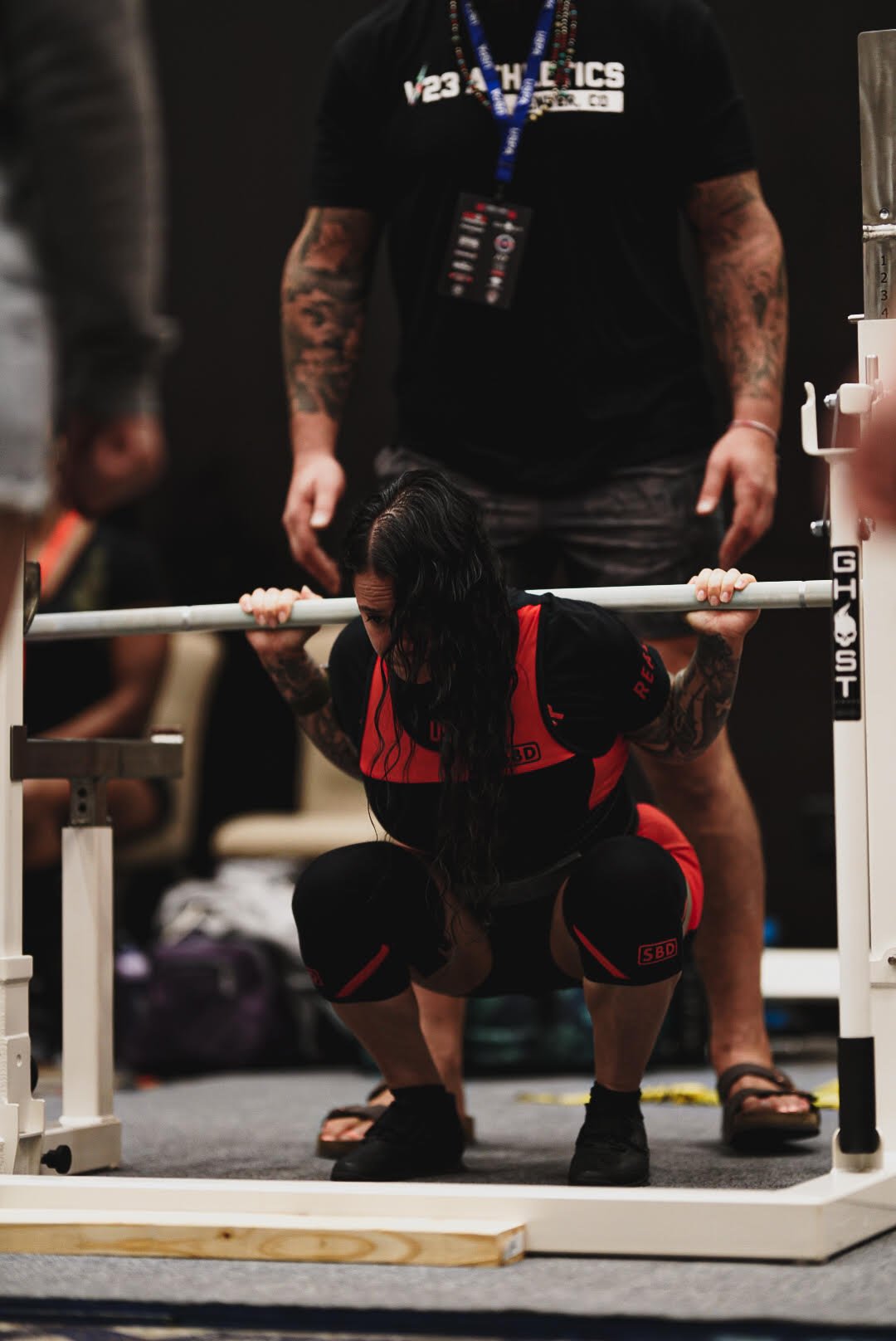 A woman in athletic gear participating in a weightlifting competition, squatting under a barbell with a spotter standing behind her in a gym or competition setting.