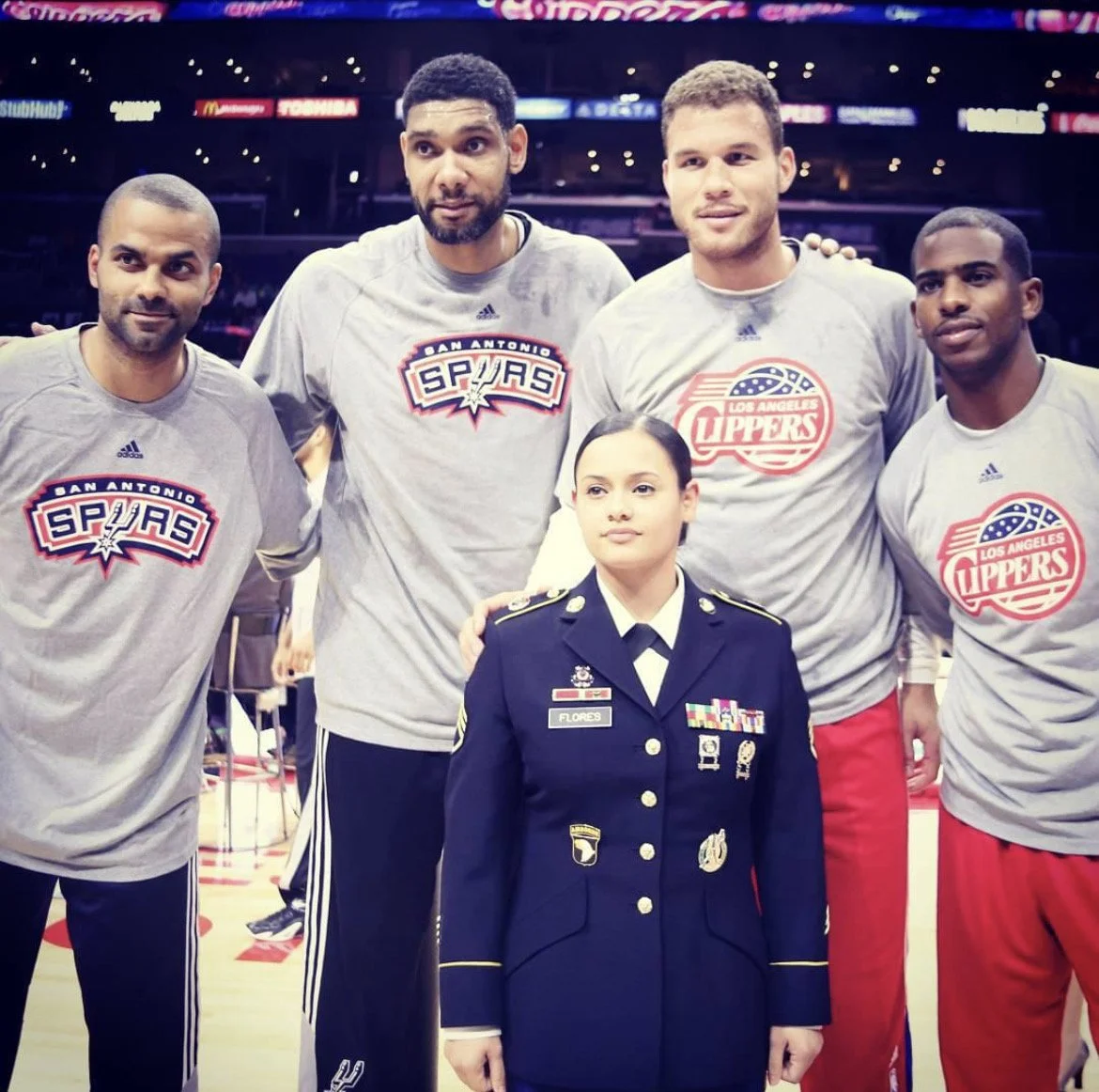 Four male basketball players wearing San Antonio Spurs and Los Angeles Clippers t-shirts pose on the court with a female military officer dressed in uniform.