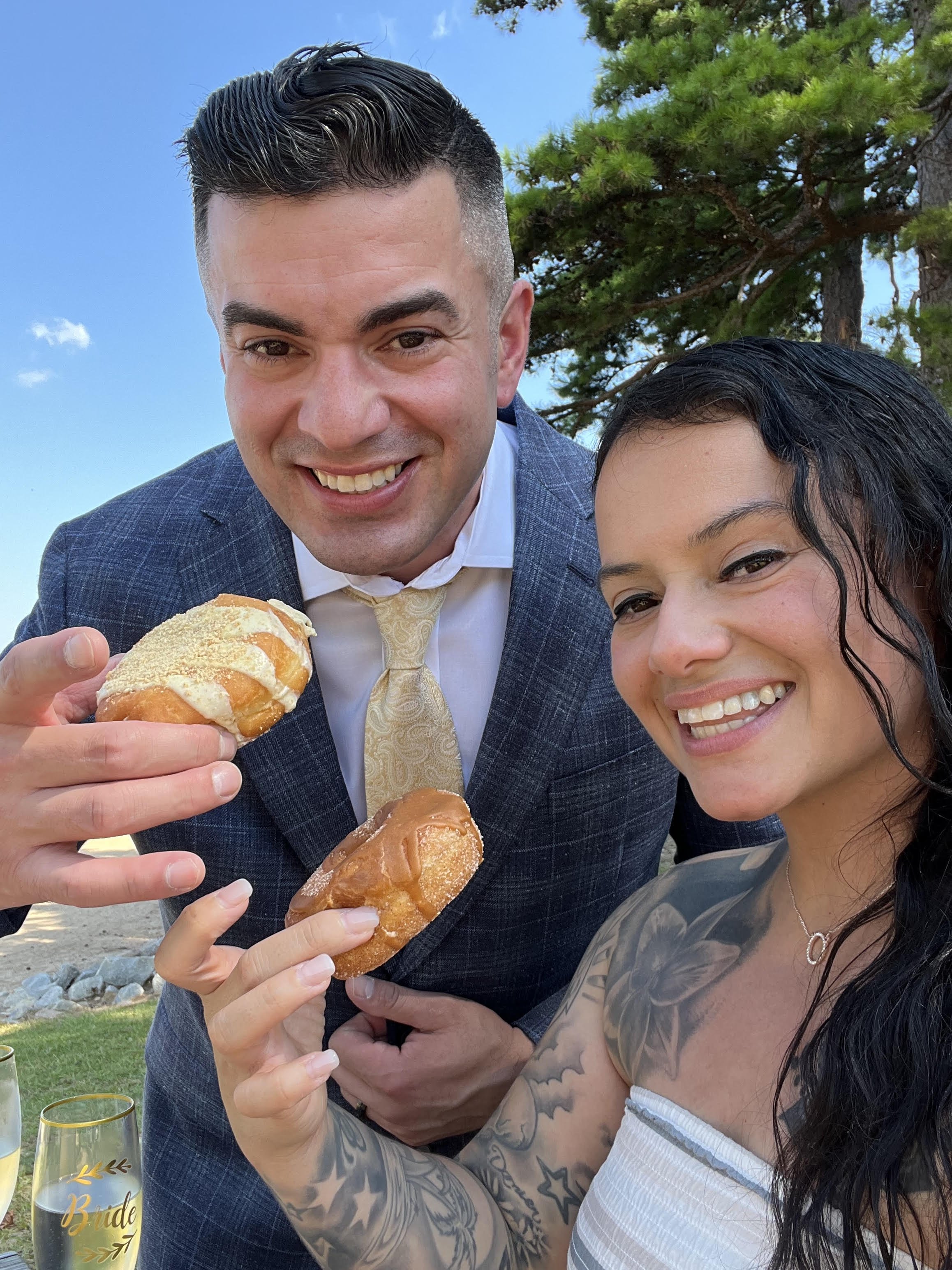 A smiling man and woman outdoors, holding food, with trees and a blue sky in the background.