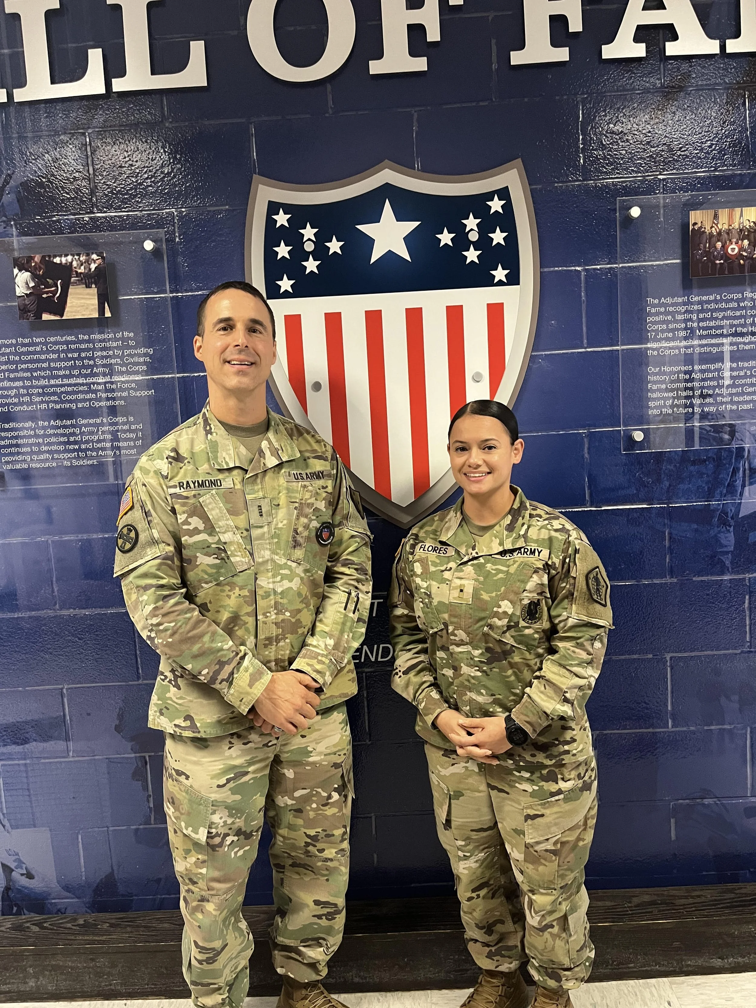 Two soldiers in camouflage uniforms standing in front of a blue wall with an insignia and informational plaques, a man on the left and a woman on the right, both smiling.