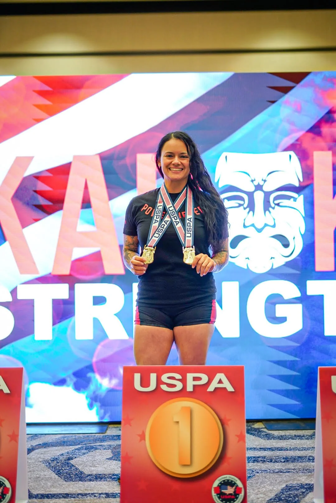 A woman standing on a podium holding two medals, with USPA branding and a large colorful background that says 'PA' and 'STRENG' behind her.