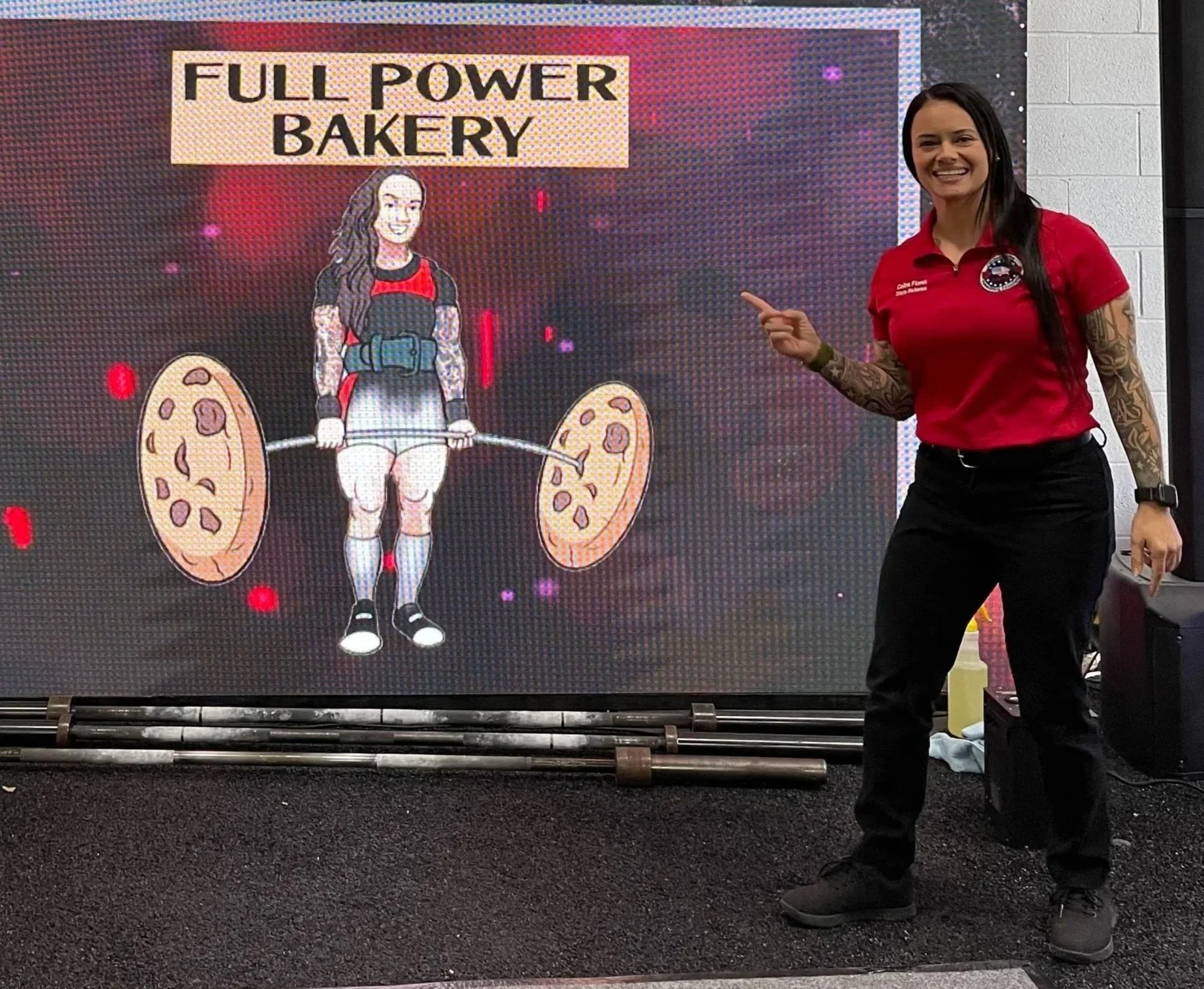 A woman with long dark hair, tattoos on her arms, wearing a red shirt and black pants, is standing and smiling while pointing at a digital display. The display features an animated illustration of a girl lifting a barbell with pizza slices on each end, and the text "FULL POWER BAKERY" at the top.
