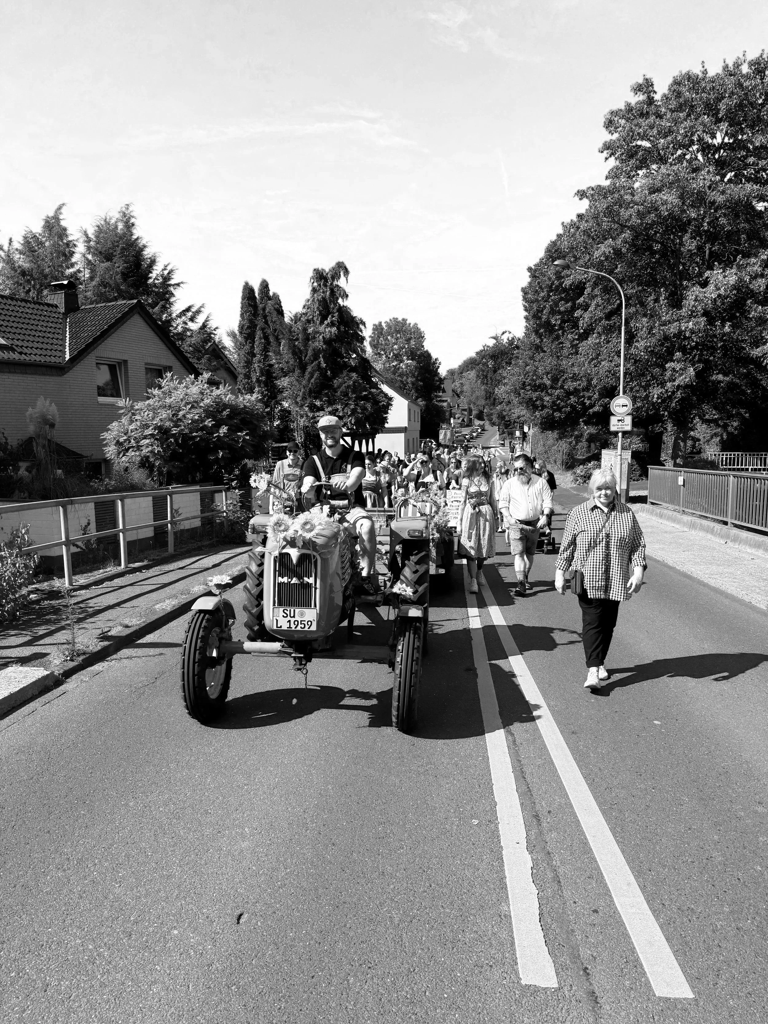 Menschen nehmen an einem Umzug oder Festzug teil, begleitet von einem grünen Traktor mit Sonnenblumen auf der Vorderseite, auf einer Ortsstraße bei schönem Wetter, umgeben von Bäumen und Häusern.