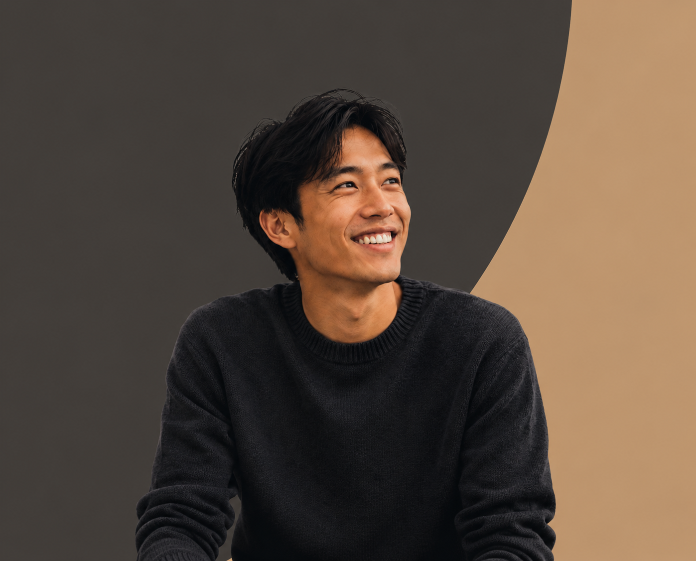Smiling young man with black hair, wearing a black sweater, sitting against a dark and beige curved background.