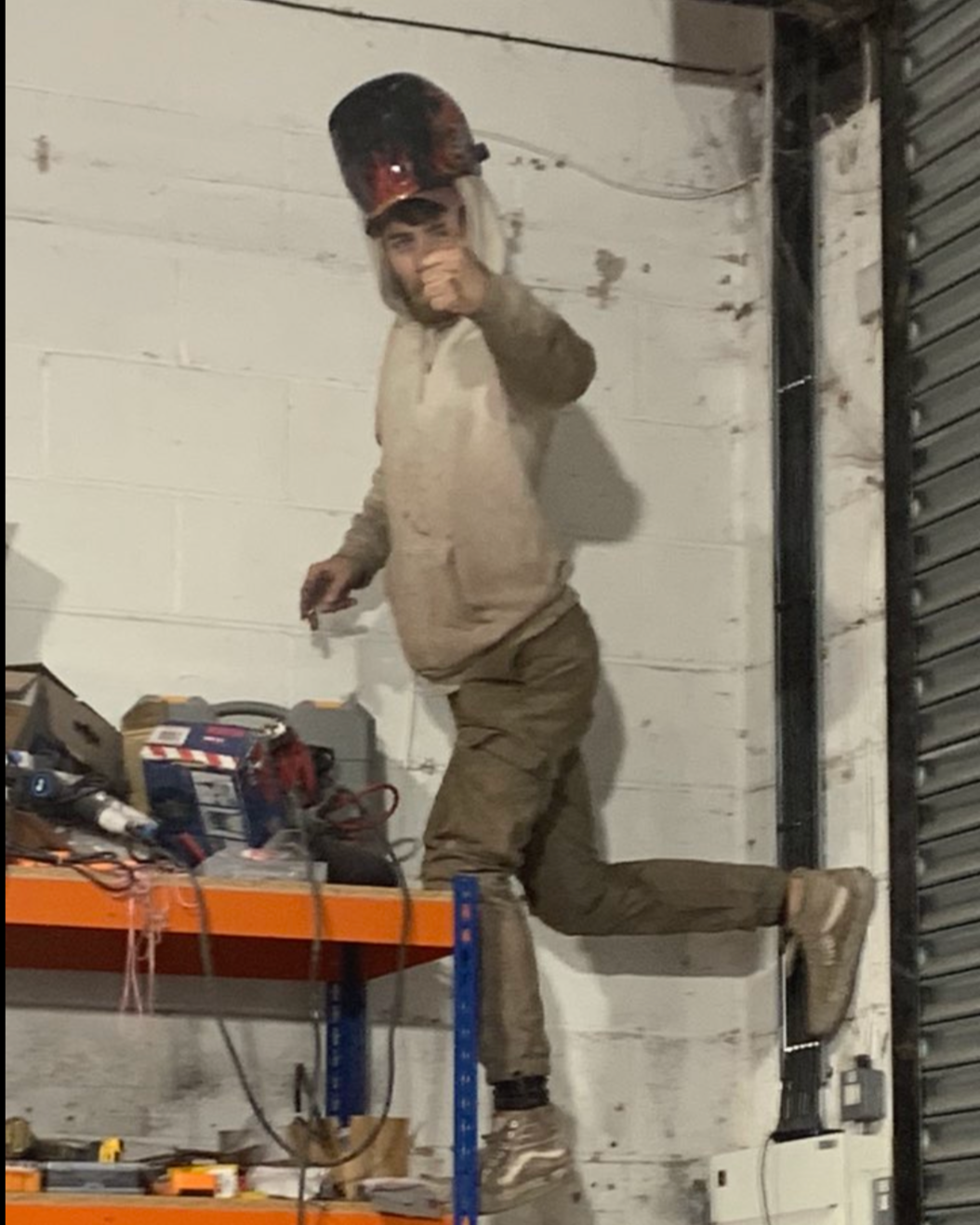 A man in work clothes and a helmet poses playfully on a metal table in a workshop or warehouse, giving a thumbs-up.