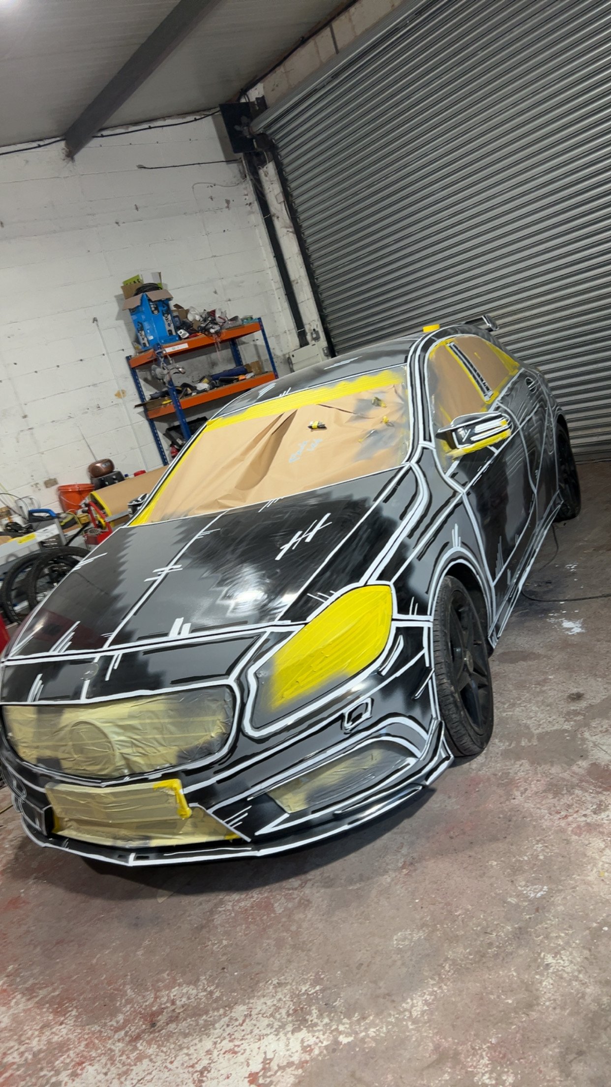Car in a garage with taped parts and masking paper, possibly undergoing paint or repair work.