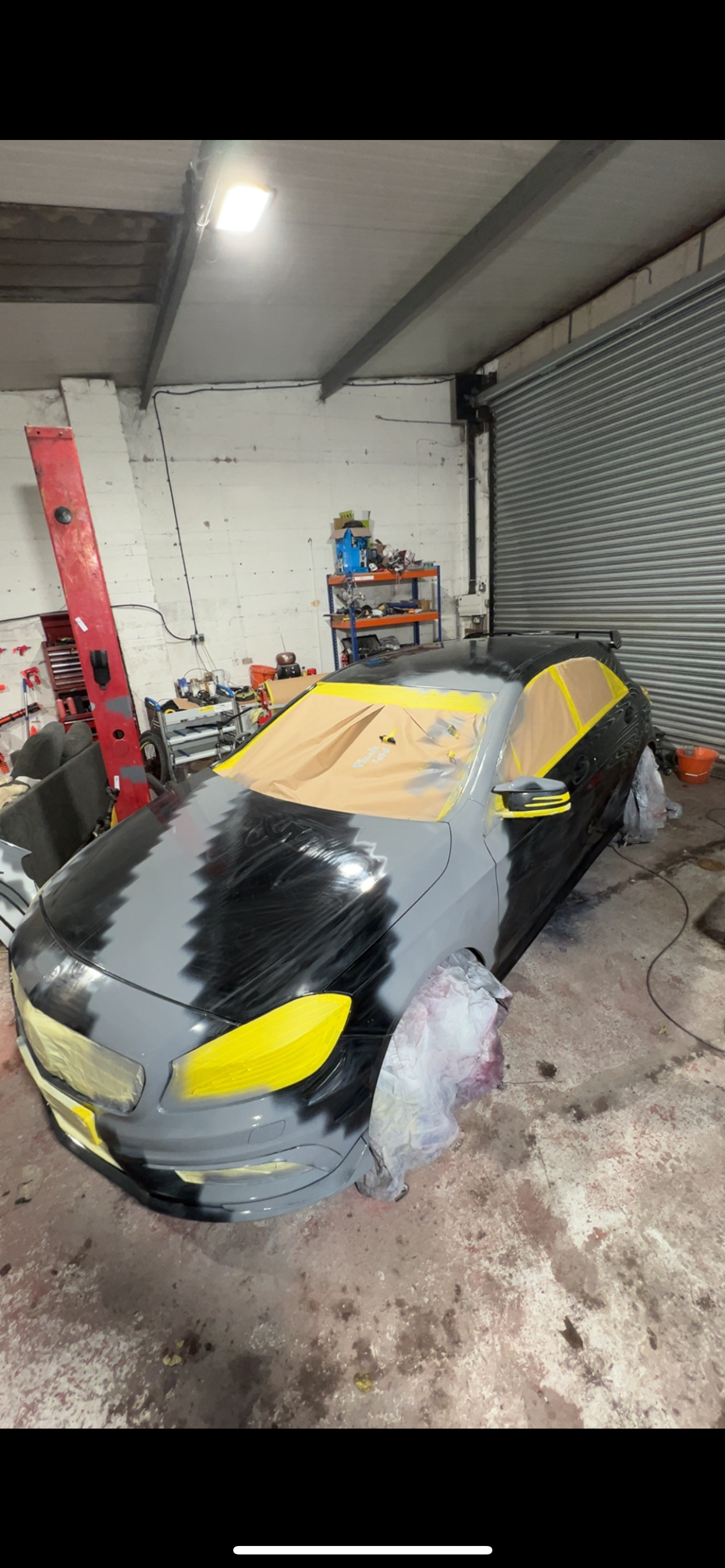 A black car in a garage being painted with black and gray paint, with yellow tape protecting specific areas. The car's front and side windows are masked with paper for painting. The garage has tools and shelves in the background.