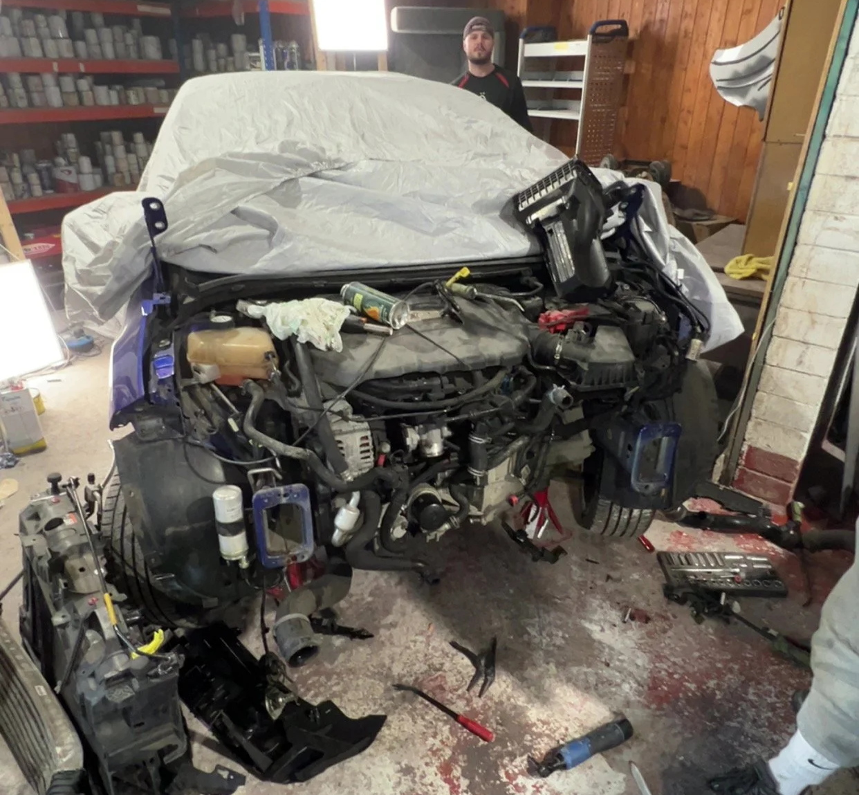 Car being repaired in a garage with tools and disassembled parts scattered on the floor, and a person in the background near shelves.