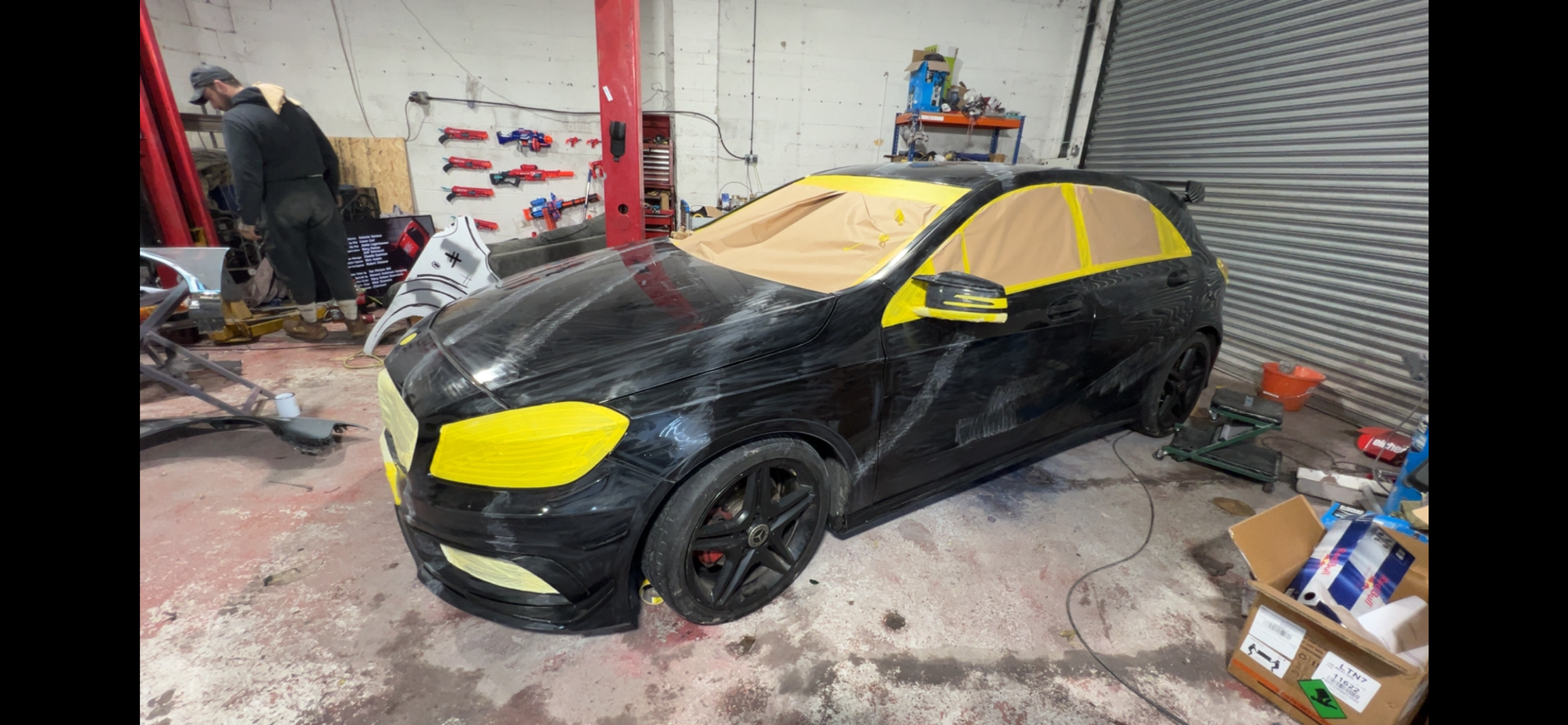 A black sports car undergoing repairs in a garage, with yellow masking tape covering its headlights, side mirrors, and windows, and a person working in the background.