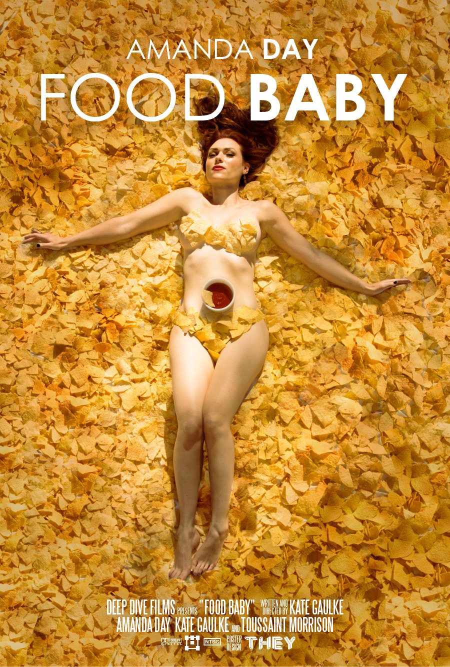 A woman lying on her back on a bed of yellow corn tortilla chips, with some chips covering her body, holding a bowl of red salsa.