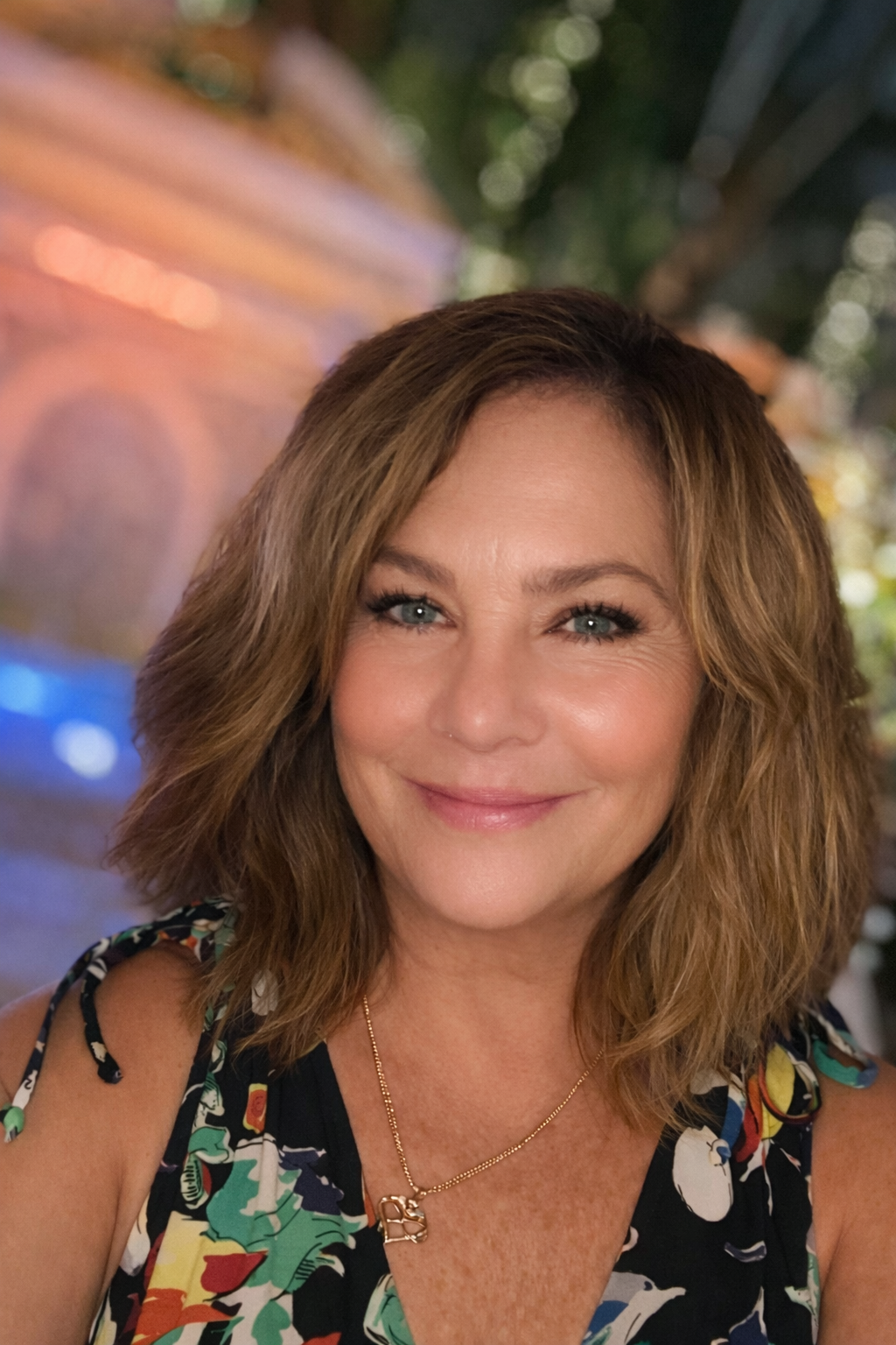 A smiling woman with shoulder-length brown hair, wearing a floral sleeveless dress and a gold necklace, at an outdoor event during the evening.