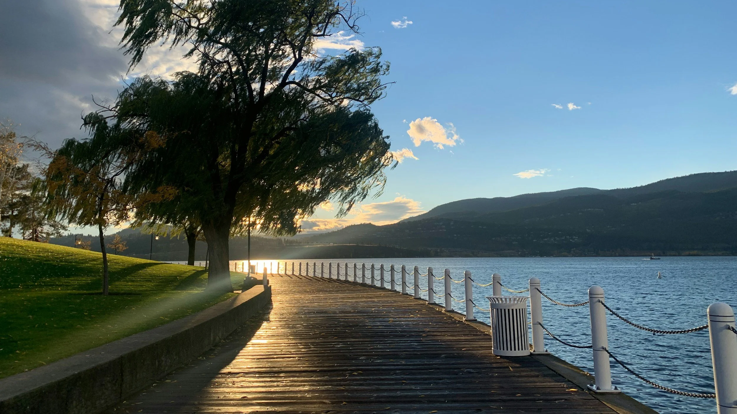 Kelowna's waterfront boardwalk at sunset with a wooden pier, a few trees on the grassy area, and mountains in the background