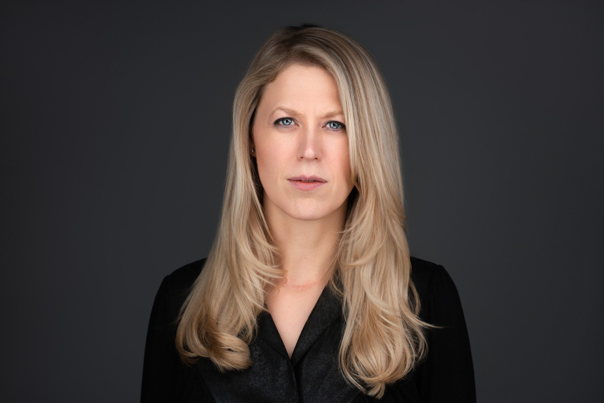A blonde woman with long wavy hair, blue eyes, and fair skin, wearing a black top, posing against a dark background.