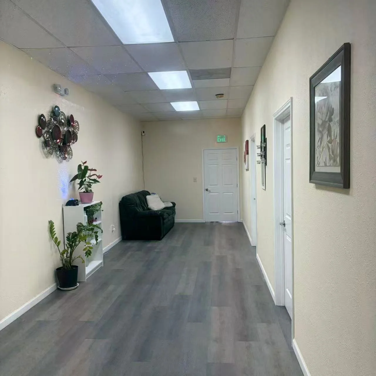 Interior hallway with yellow walls, gray flooring, white ceiling tiles, a black sofa with pillows, framed pictures, and potted plants.