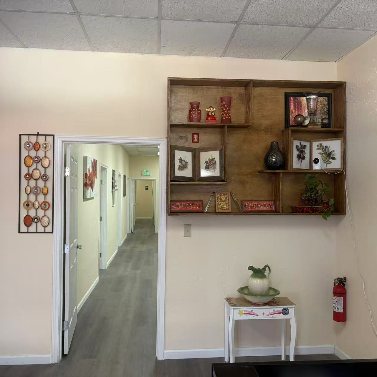 Interior view of a hallway with walls decorated with artwork and a wooden shelf with decorative vases, framed botanical prints, and a plant. There is a small table with a ceramic pitcher and a fire extinguisher beside it.