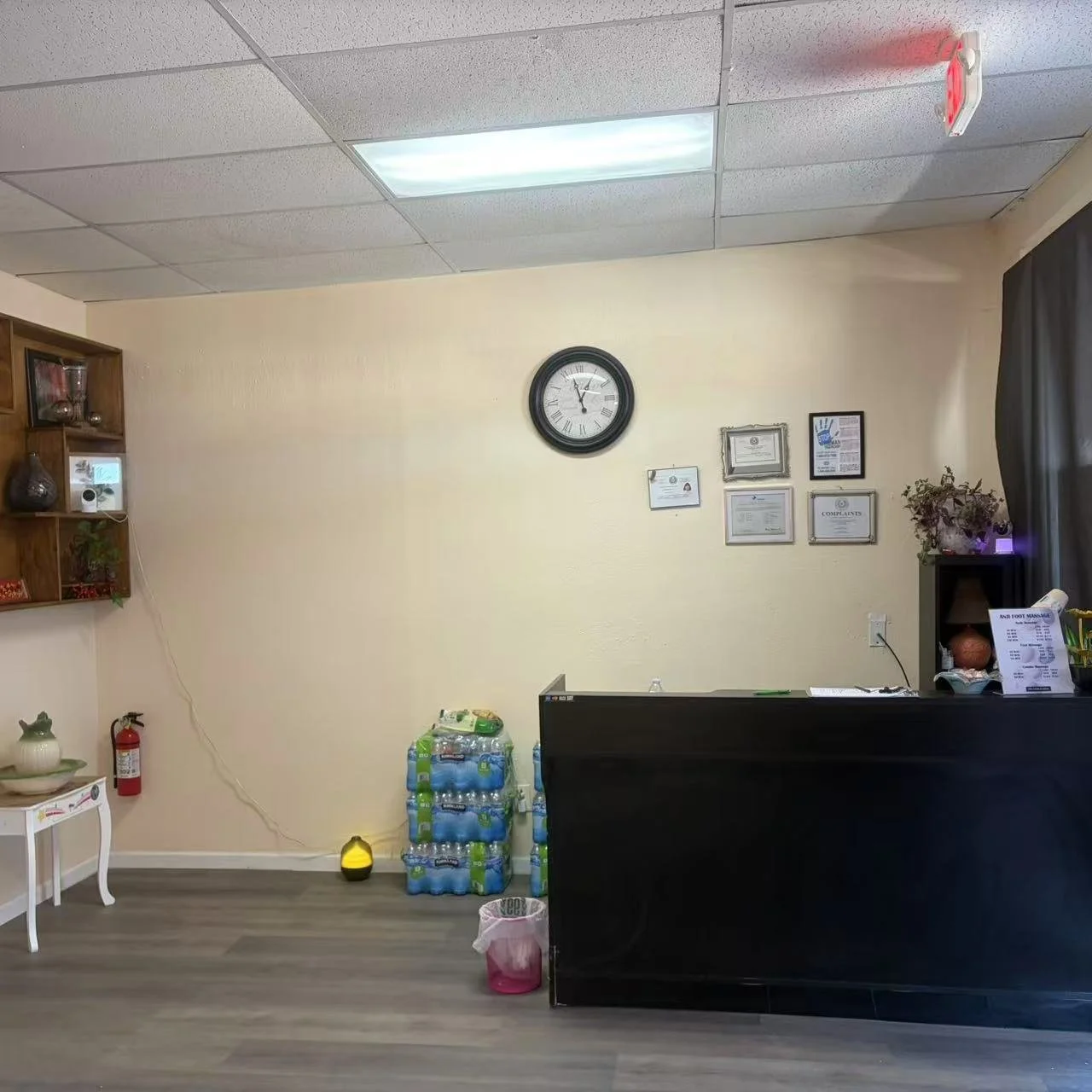 Interior of an office or reception area with a clock on the wall, certificates on display, a black desk, bottles of water, a fire extinguisher, and decorative items on shelves and tables.