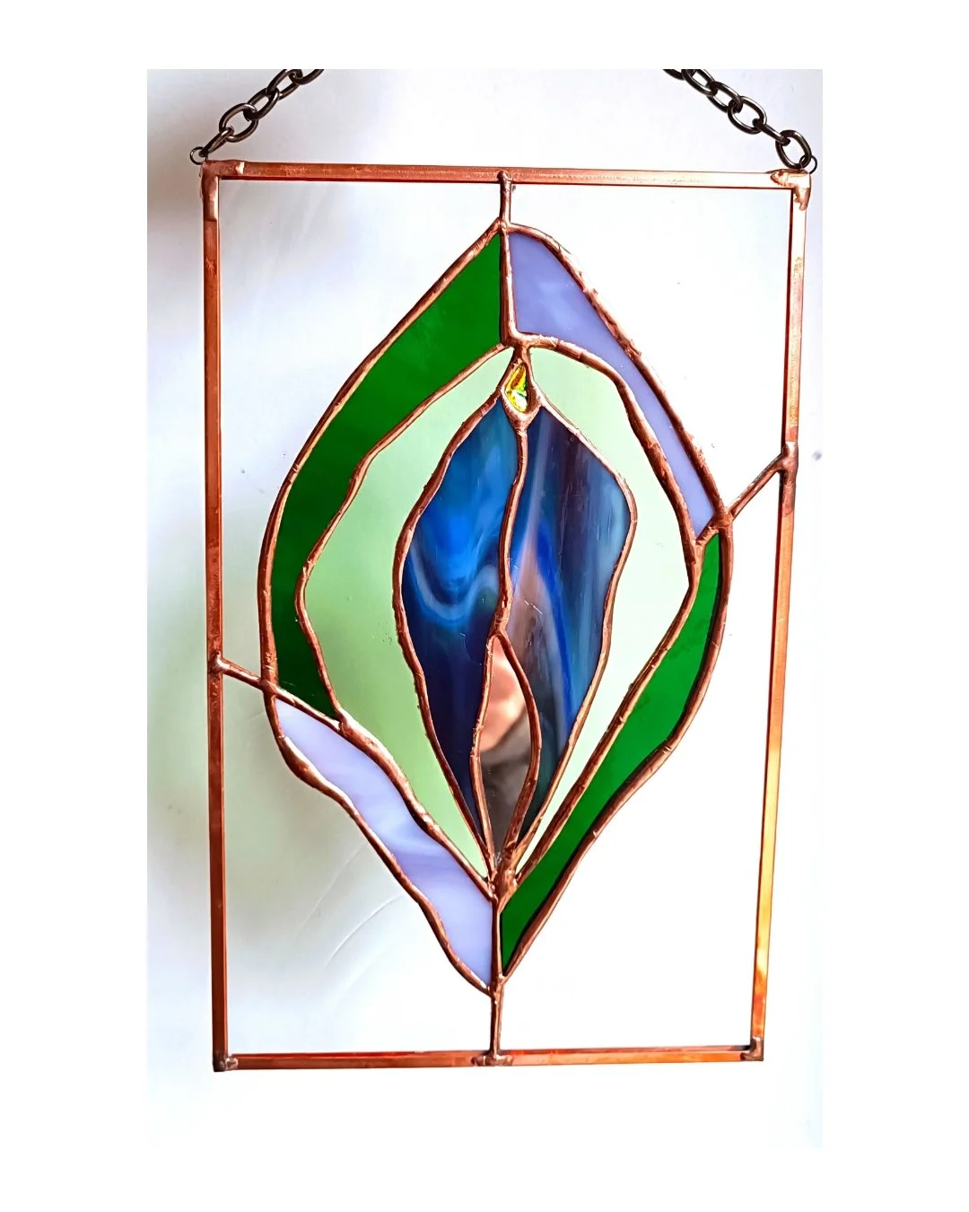 Vitrail coloré avec des formes abstraites en vert, bleu et violet, dans un cadre en cuivre, suspendu par une chaine.