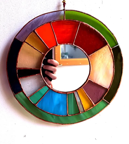 Un miroir rond en vitrail coloré accroché au mur, avec une personne qui prend la photo en se reflétant dans le miroir. c'est Diane qui vous accueil dans son bel atelier à Lille.