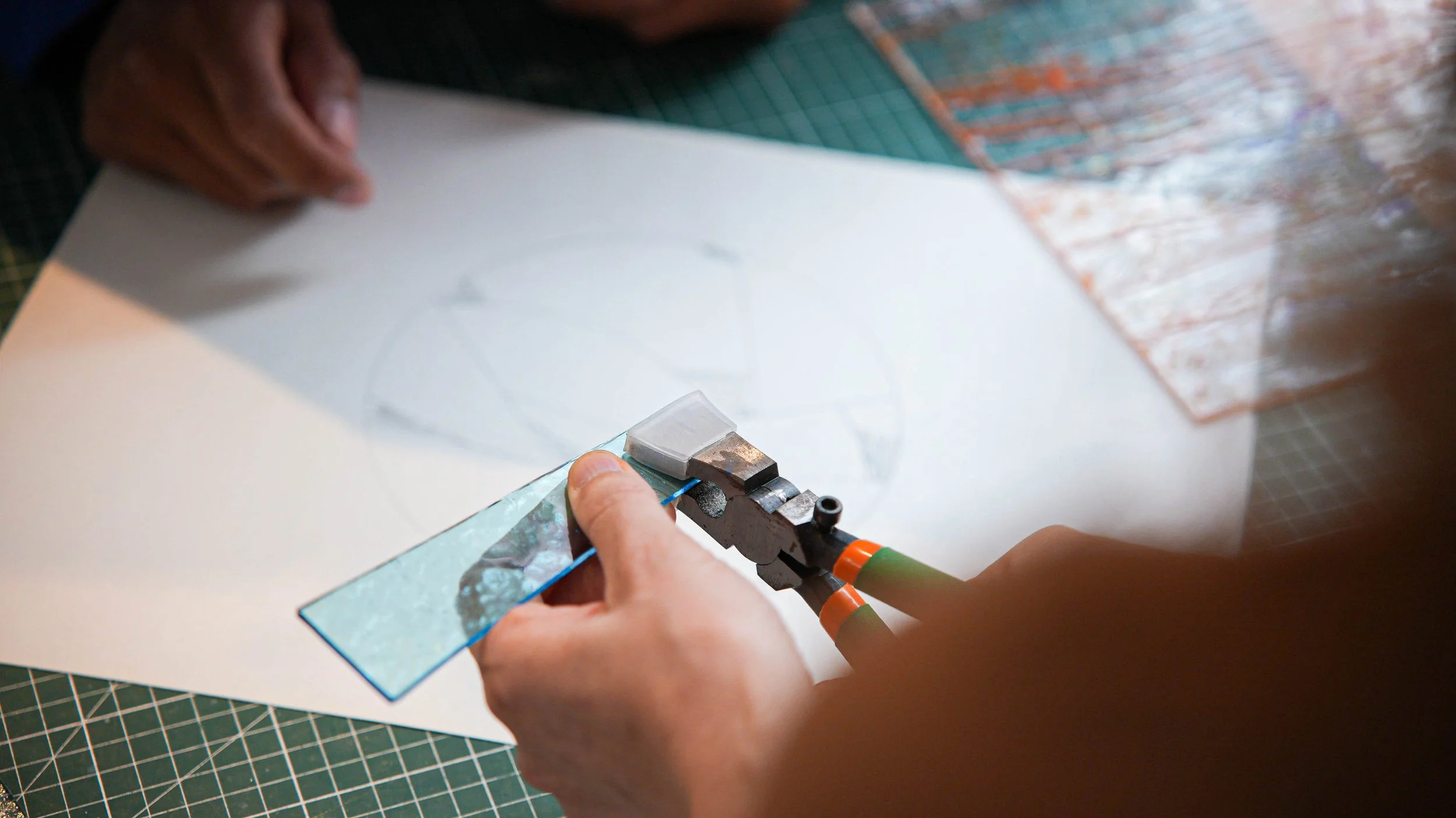 Une personne utilise une pince pour couper un morceau de verre sur un tapis de découpe. Dans le fond, un papier blanc avec un dessin partiellement visible et une grille de découpe.