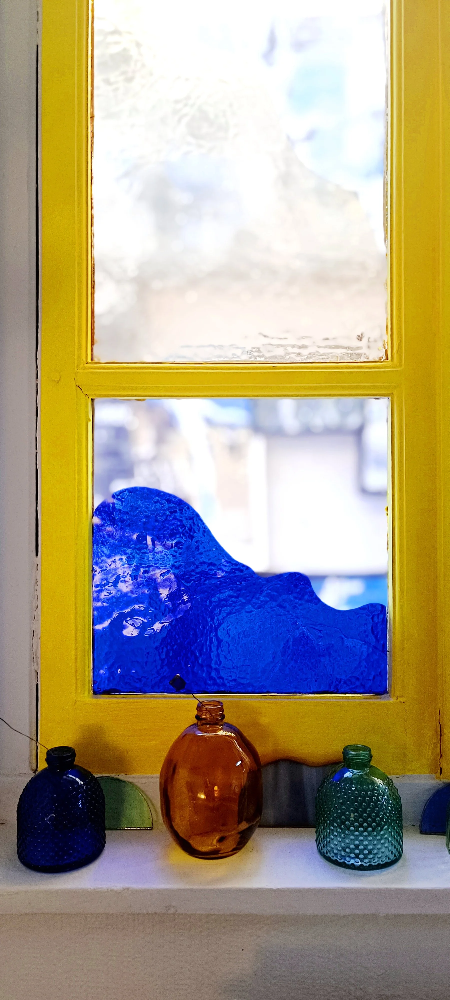 Vase en verre orange sur une étagère avec deux autres vases en verre bleu et vert, devant une fenêtre jaune avec des vitraux. Des fleurs en verre sont dans les petites vases colorés en verre. La fenêtre a des pièces de verres colorés aux fenêtres.
