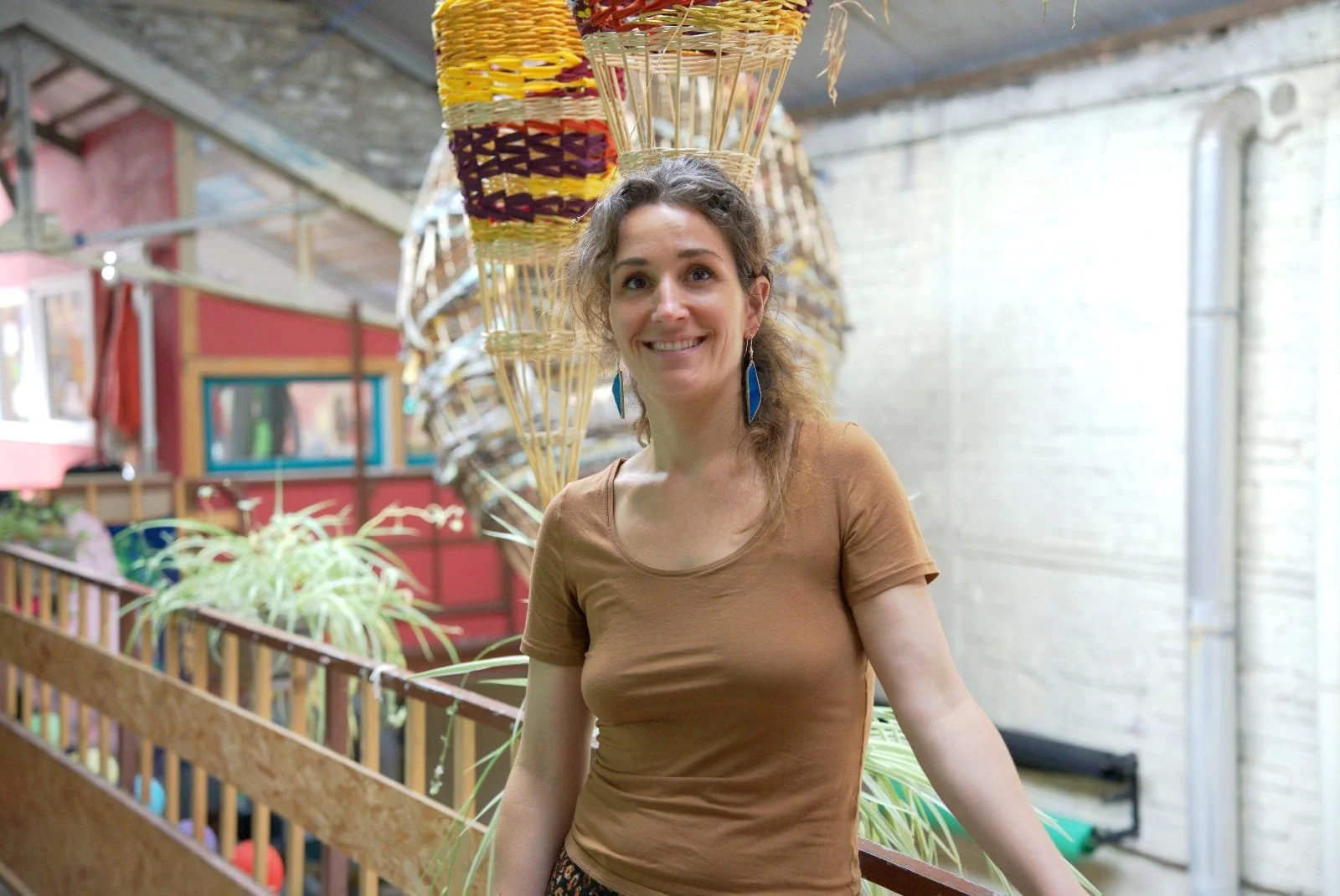 Une femme souriante avec des vêtements marron et des boucles d'oreilles bleues en vitrail, dans un lieu coloré avec des décorations en osier et des plantes vertes. C'est là que Diane vous accueil pour un rendez-vous dans son atelier.