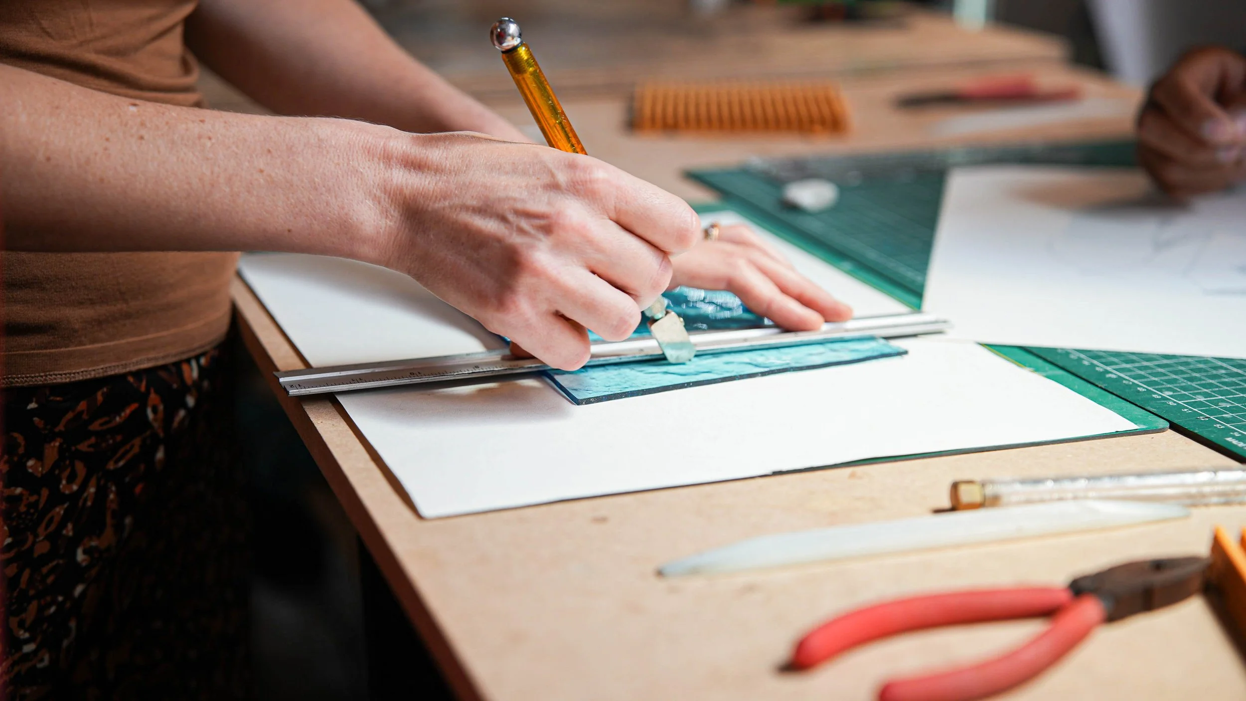 Une personne utilisant un cutter pour découper un motif en verre sur une planche de travail, entourée d'outils de travail, avec un focus sur les mains et les outils.