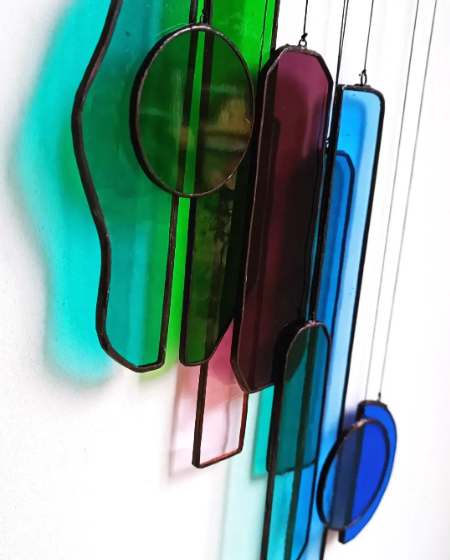 Description d'un mobile en verre coloré suspendu, inclut des formes ovales et rectangulaires en verre bleu, vert, rouge et transparent, entourées de lignes métalliques, mobile en vitrail moderne de la vitrailliste Diane Frank.