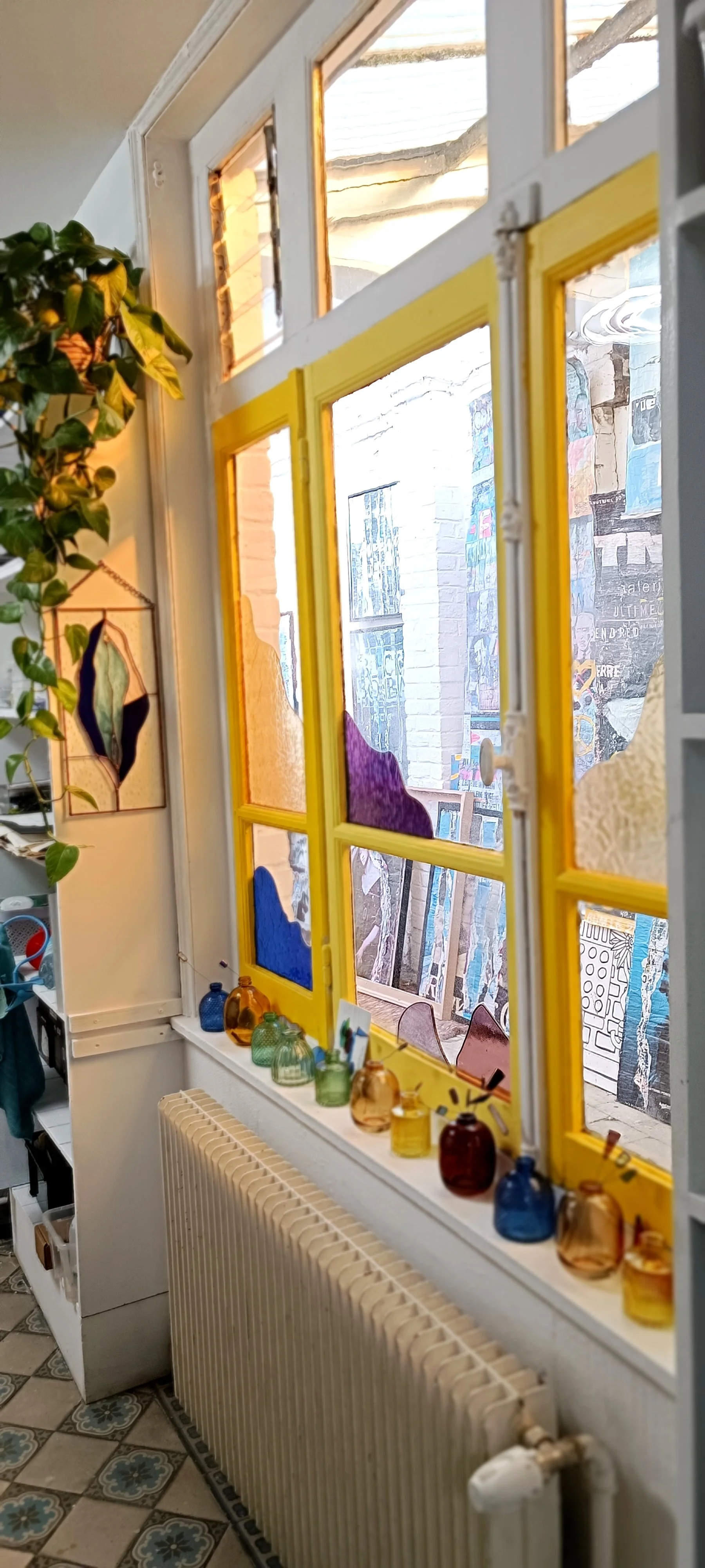 Fenêtre avec cadre jaune, décorée avec des petits vases colorés sur le rebord, vue extérieure d'un mur avec affiches et œuvres artistiques, intérieur avec plantes et objets décoratifs. intérieur de l'atelier Le vitrail de Diane, artiste vitrailliste.
